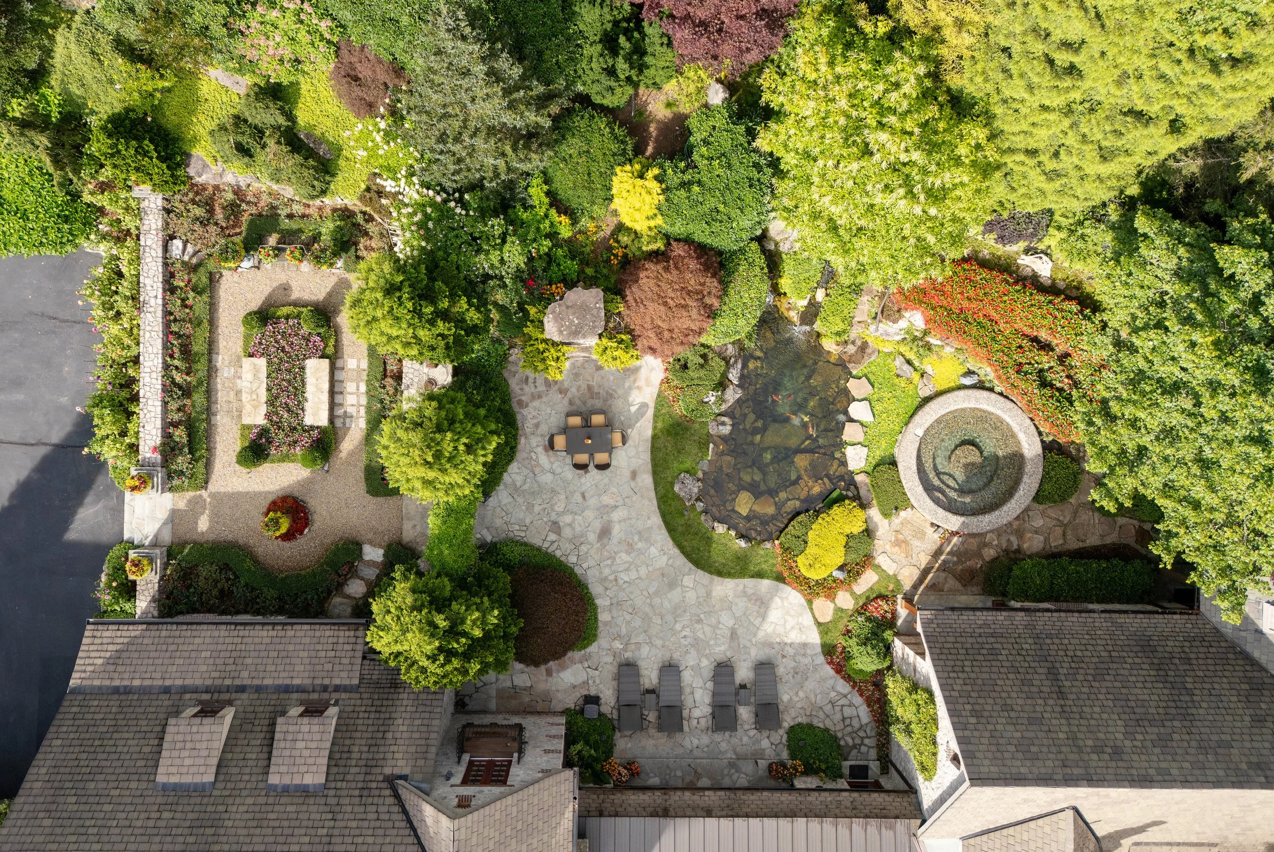 An aerial view of a landscaped backyard with various trees, shrubs, flower beds, a small pond, a stone patio with a table and chairs, and a surrounding stone and hedge border.