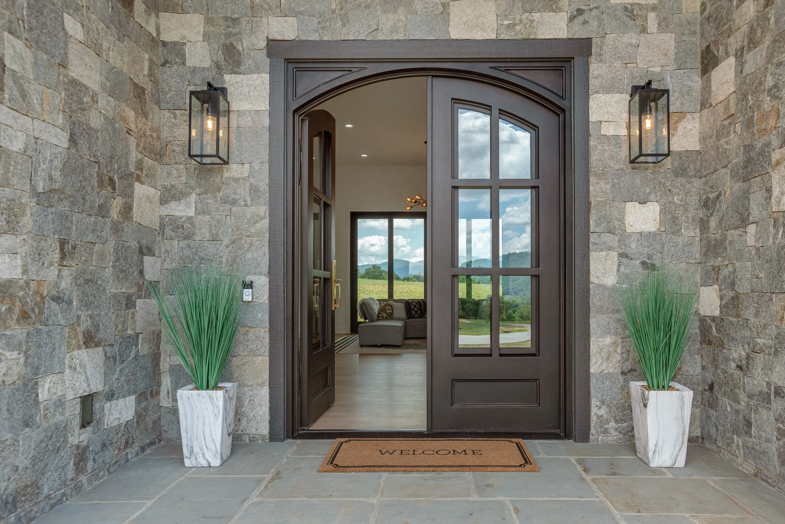 Entryway with stone walls, a dark wooden door, glass panels, and potted plants flanking the door, leading into a living room with a view of mountains and blue sky.