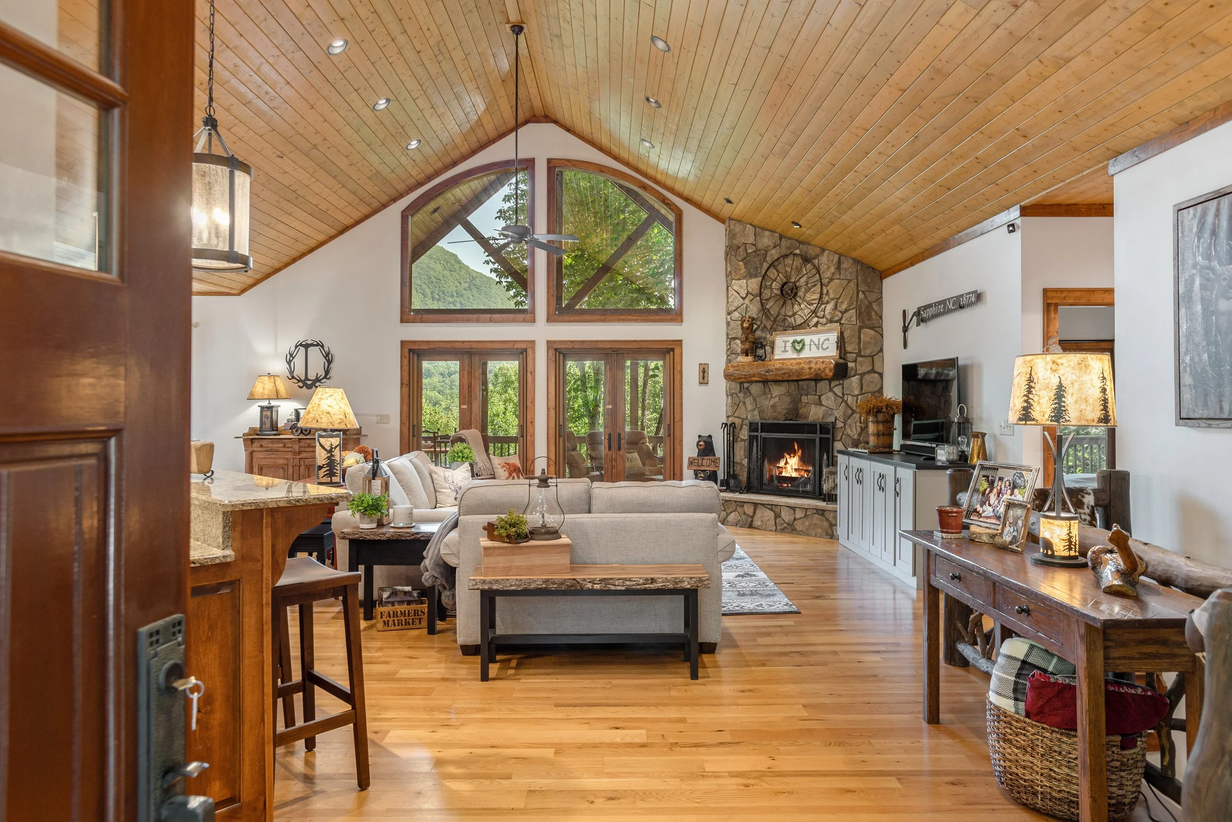 Living room with a vaulted wooden ceiling, large windows, and a stone fireplace. Furniture includes sofas, a coffee table, and lamps. Decor features signs, framed photos, and a ceiling fan.