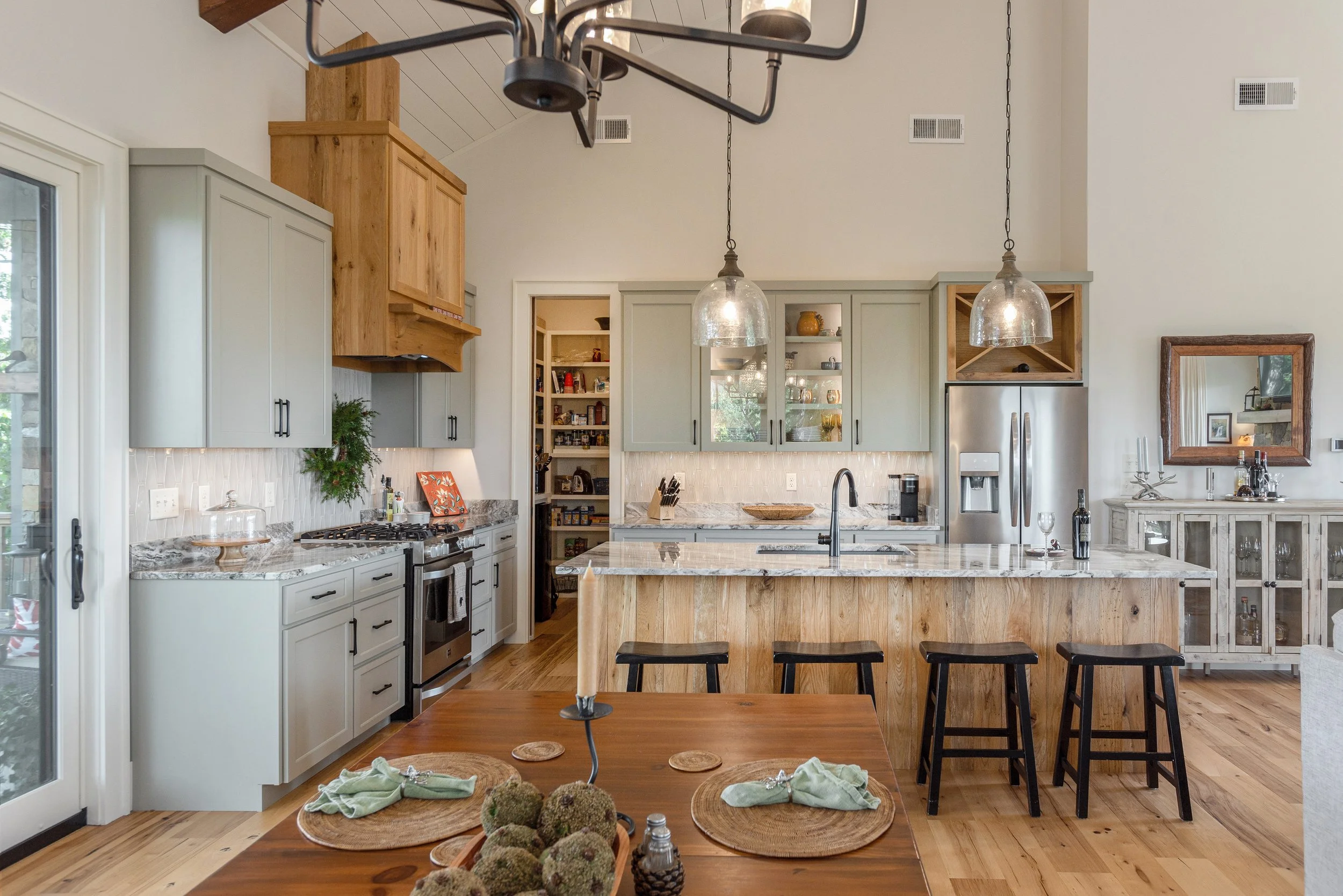 A modern kitchen with white and light gray cabinetry, a marble island, and pendant lighting. There is a stove, fridge, and open pantry, with a dining table set with placemats and napkins.