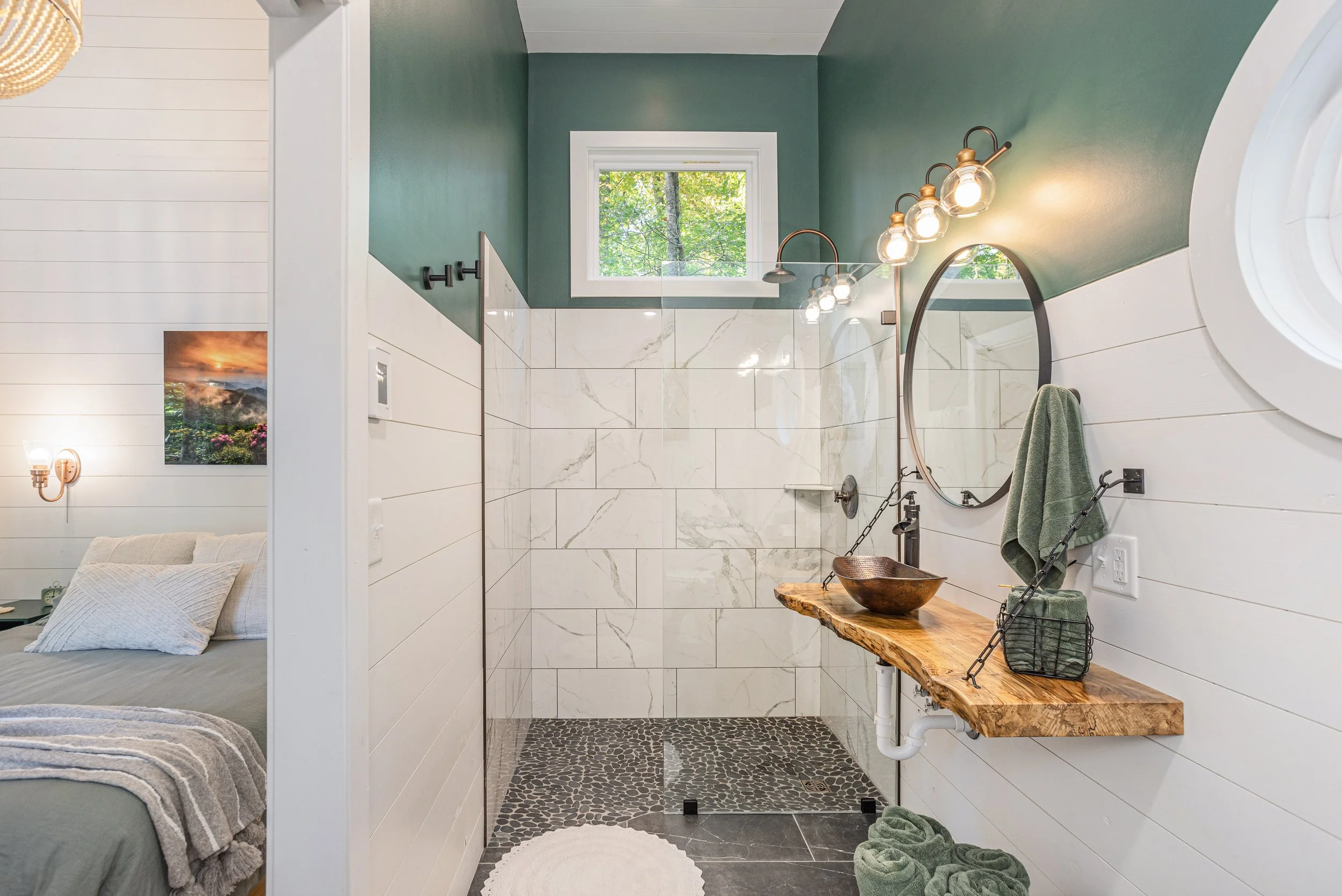 Interior view of a modern bathroom with a green ceiling, white tiled walls, a window showing trees outside, and a pebble-patterned floor. There are four exposed bulb vanity lights, a round mirror, a towel hanging on a hook, and a wooden bench with a 