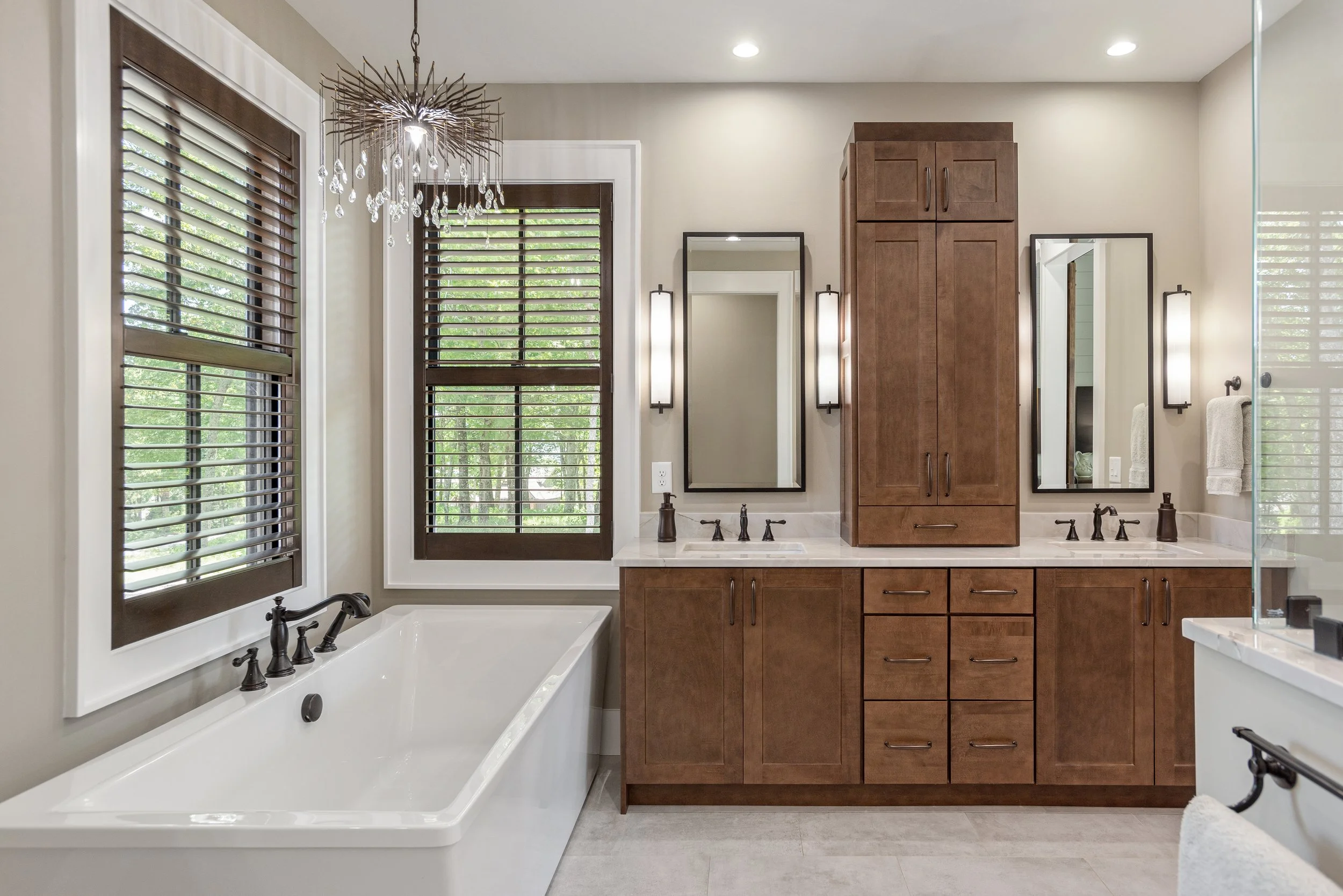 Modern bathroom with a white bathtub, dark wood cabinets, and two windows with brown blinds. There are two mirrors and wall-mounted lights next to the cabinet, and a chandelier hanging from the ceiling.