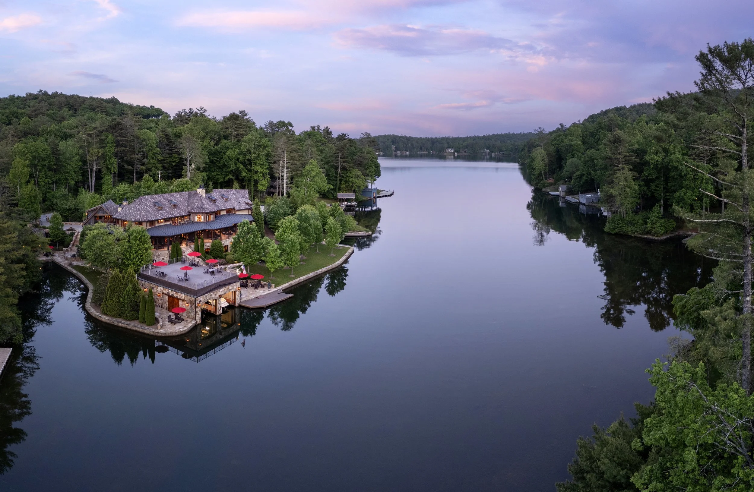 A large house with a deck and outdoor seating area, situated on the edge of a calm lake surrounded by lush green trees, with a sky showing soft pastel colors at sunset.