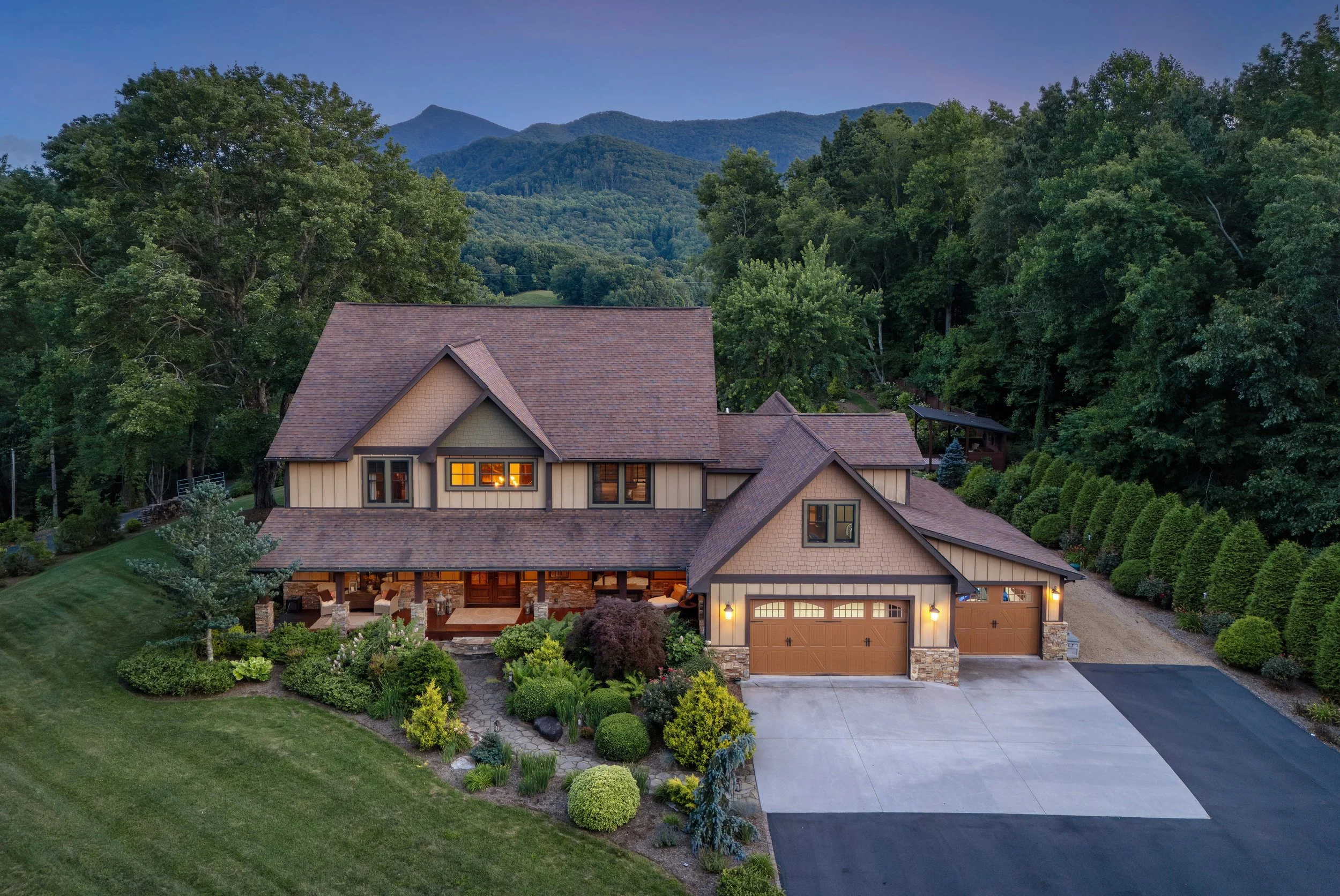 A house with a brown roof and beige walls, surrounded by a well-maintained garden, with mountains and trees in the background during dusk.