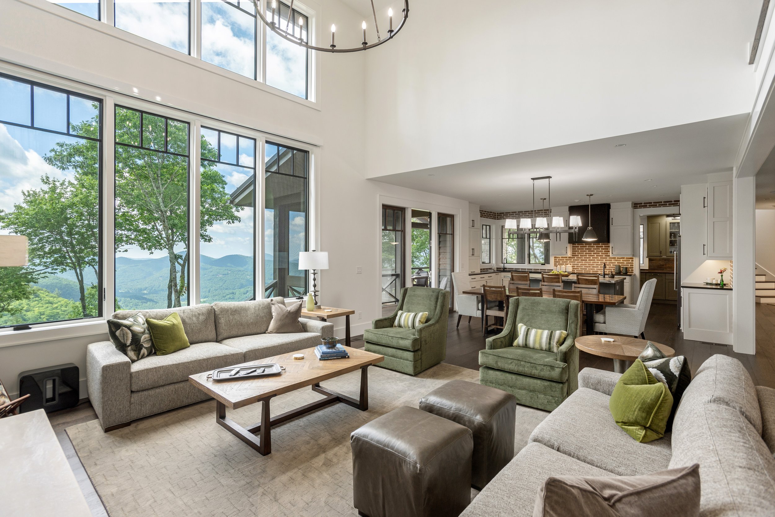 Open living room with large windows revealing a mountain view, featuring multiple sofas, green armchairs, wooden coffee tables, a rug, and a chandelier hanging from the ceiling.