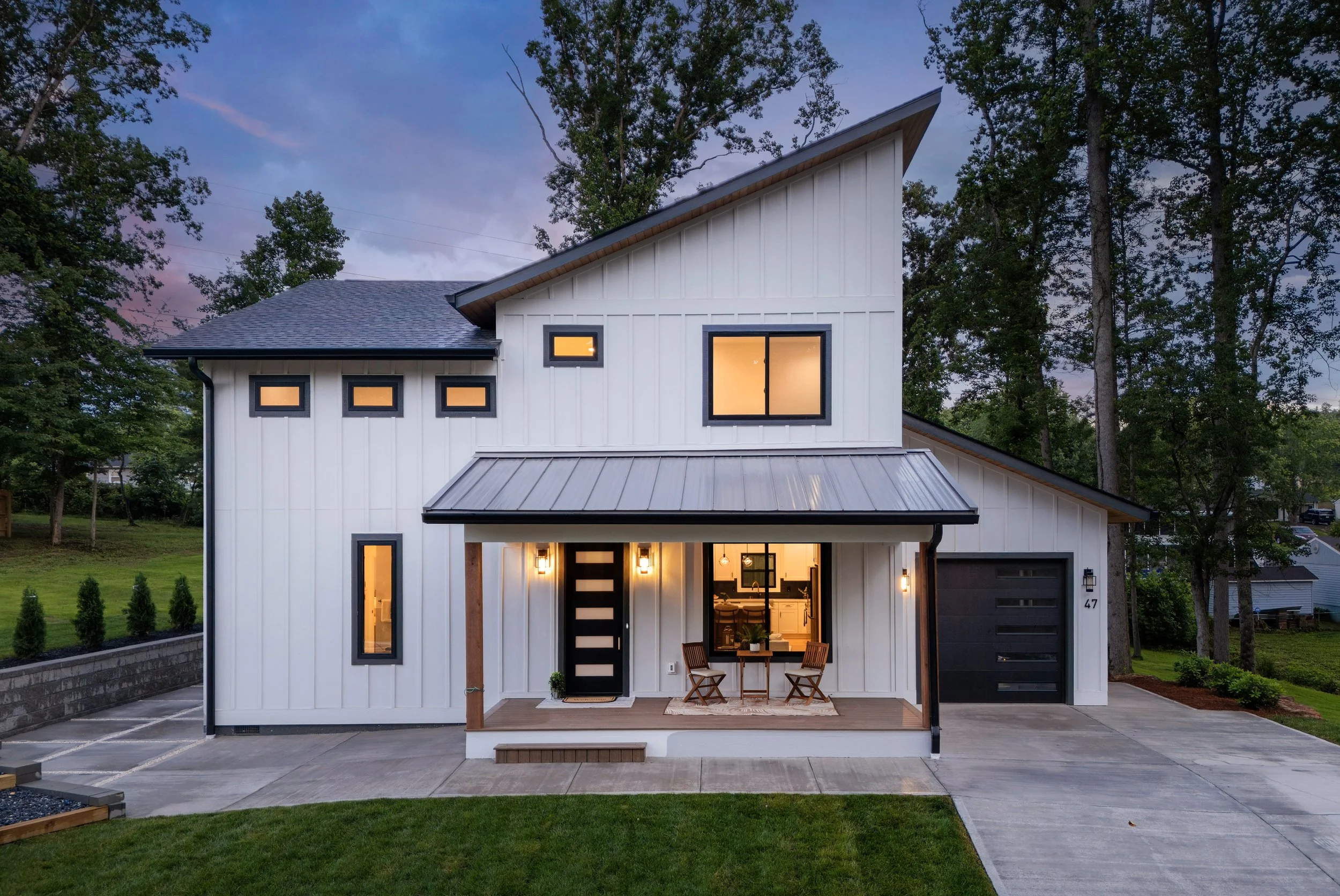 Modern white house with black window frames, front porch with chairs, and a garage, surrounded by trees and a parking lot, during evening with interior lights glowing.