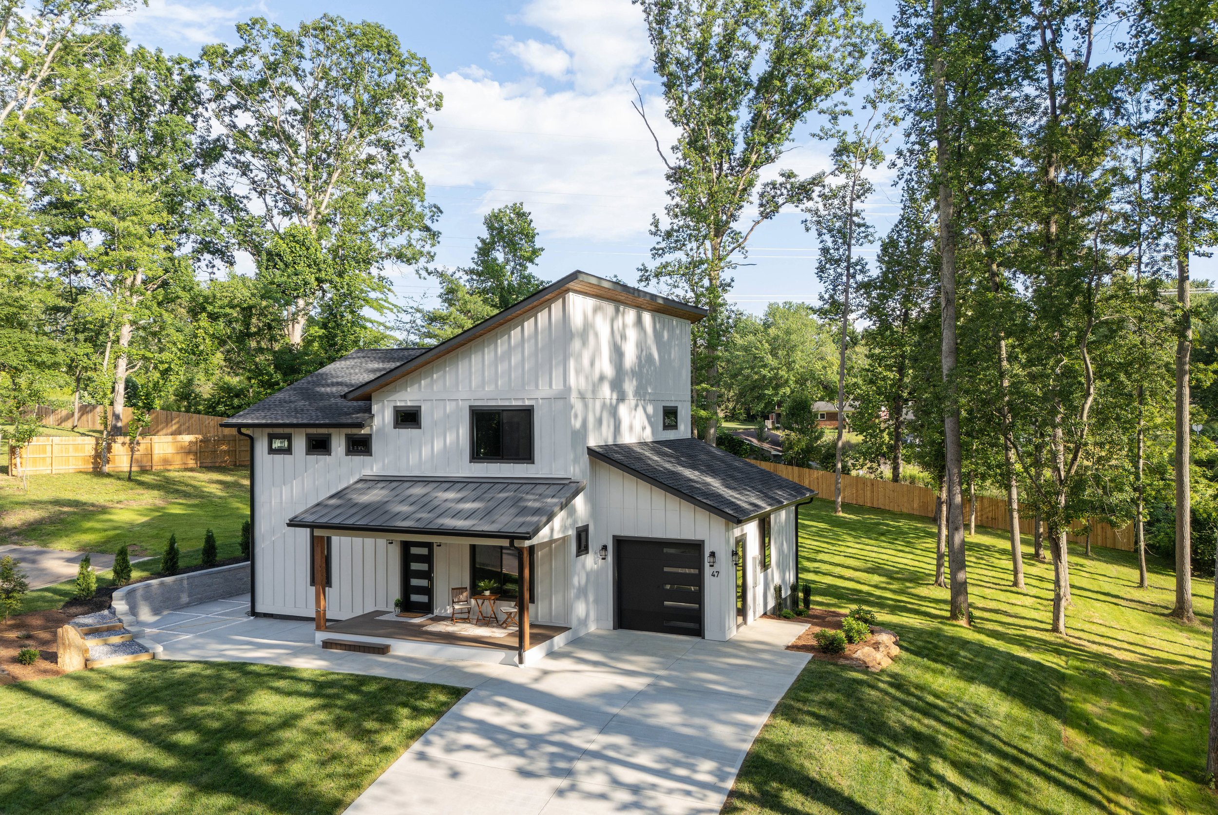 Front view of a modern white house with black garage door, small porch with seating, surrounded by green lawn and tall trees, under a partly cloudy sky.