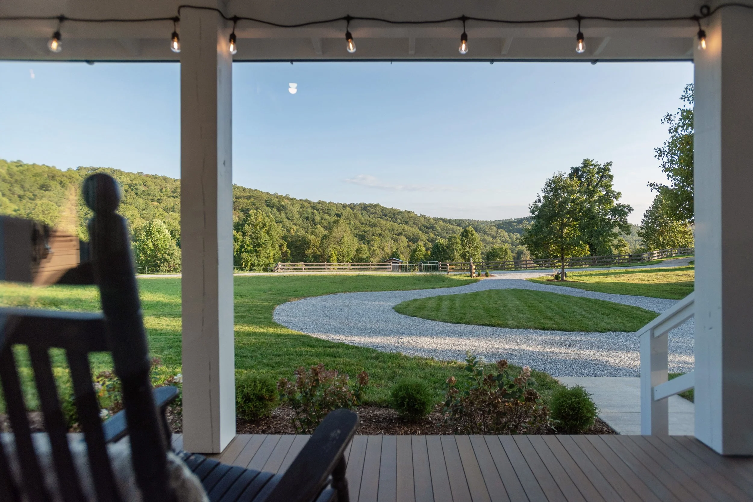 View from a porch showing a garden with a curved gravel pathway, a green lawn, trees, a fenced area, and a small shed, with hills covered in trees in the background under a clear sky.