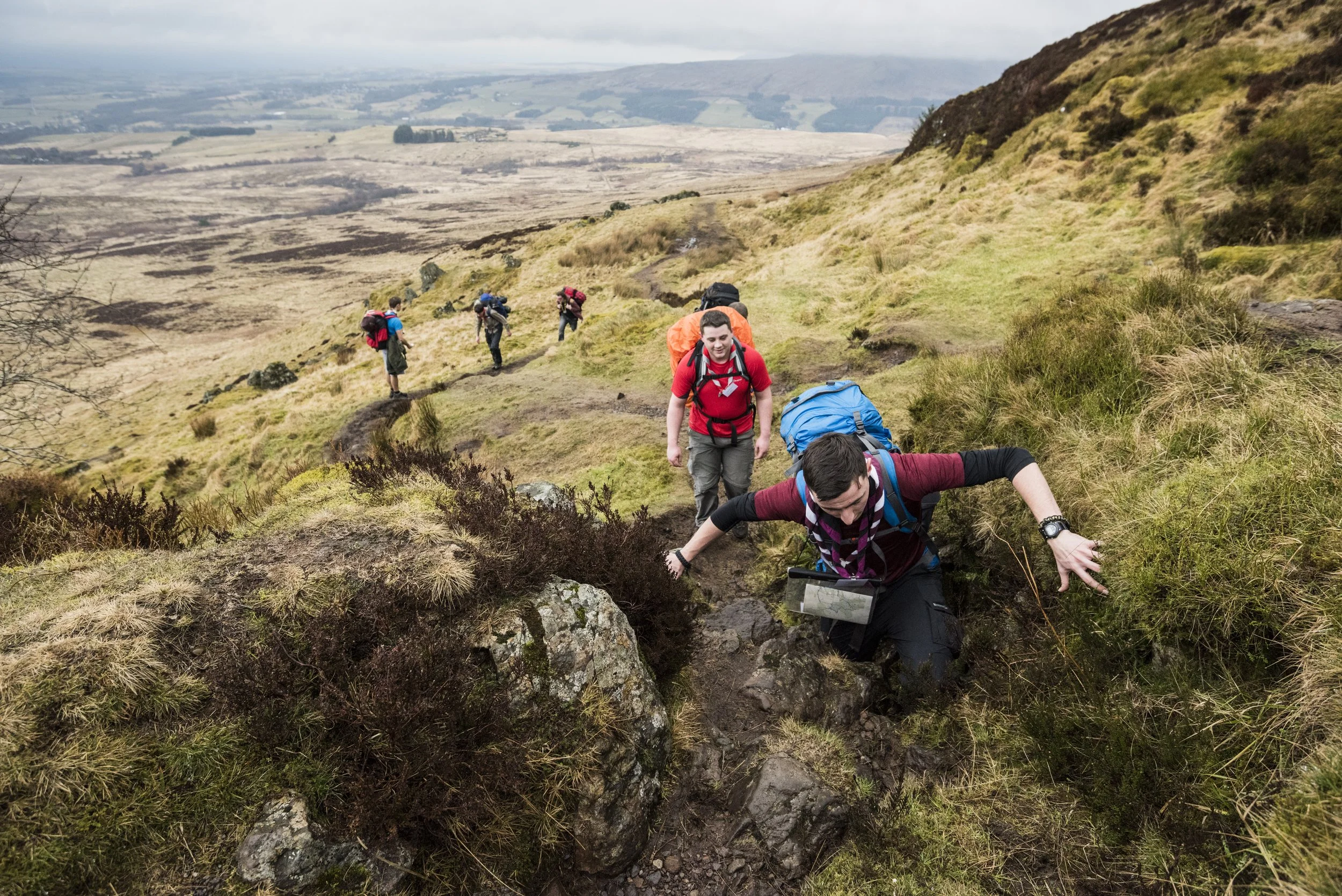 Group of hikers climbing a grassy hillside with rocky terrain in a mountainous landscape.