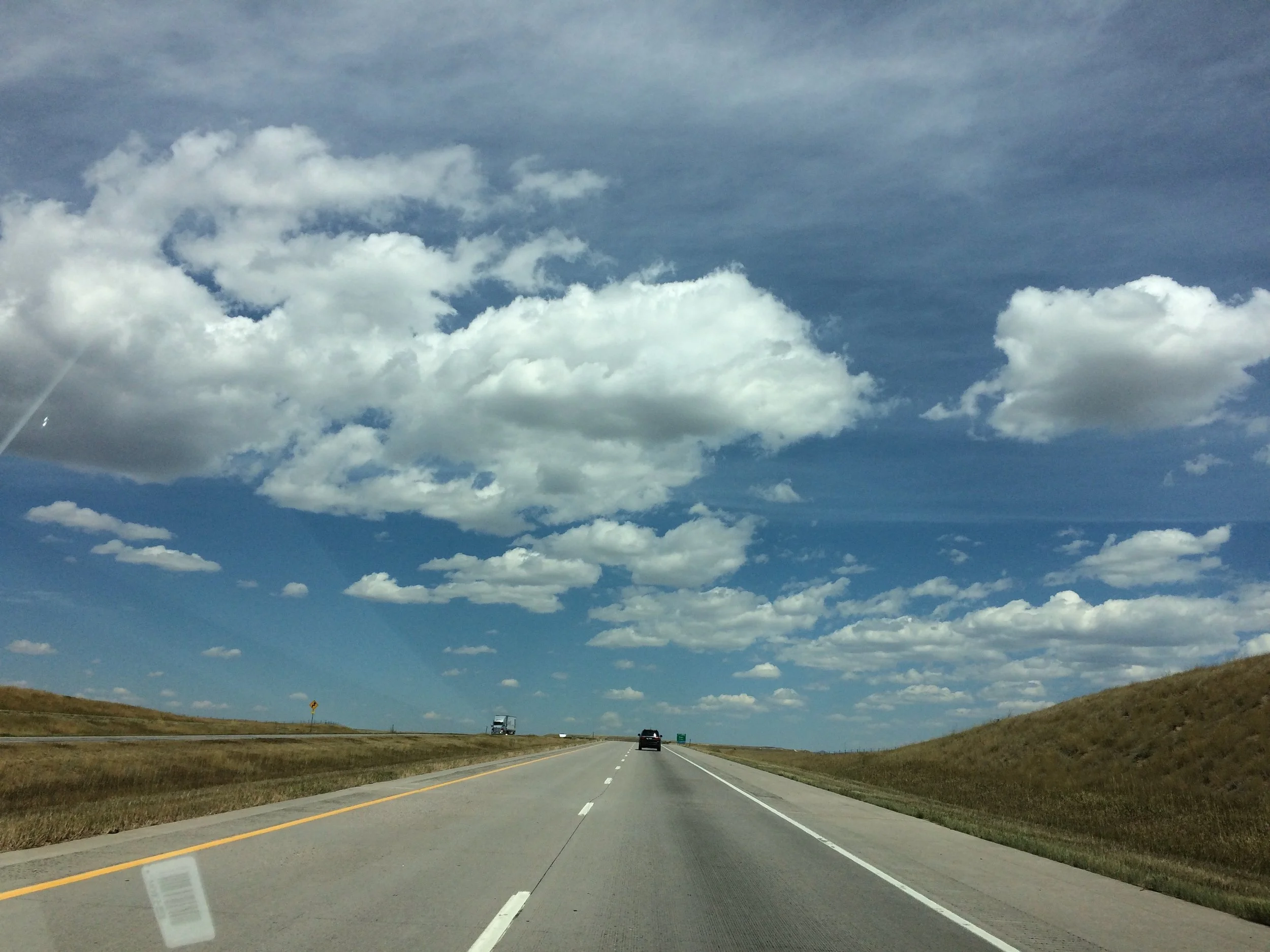 An open highway under a blue sky with scattered white clouds, surrounded by grassy hills.