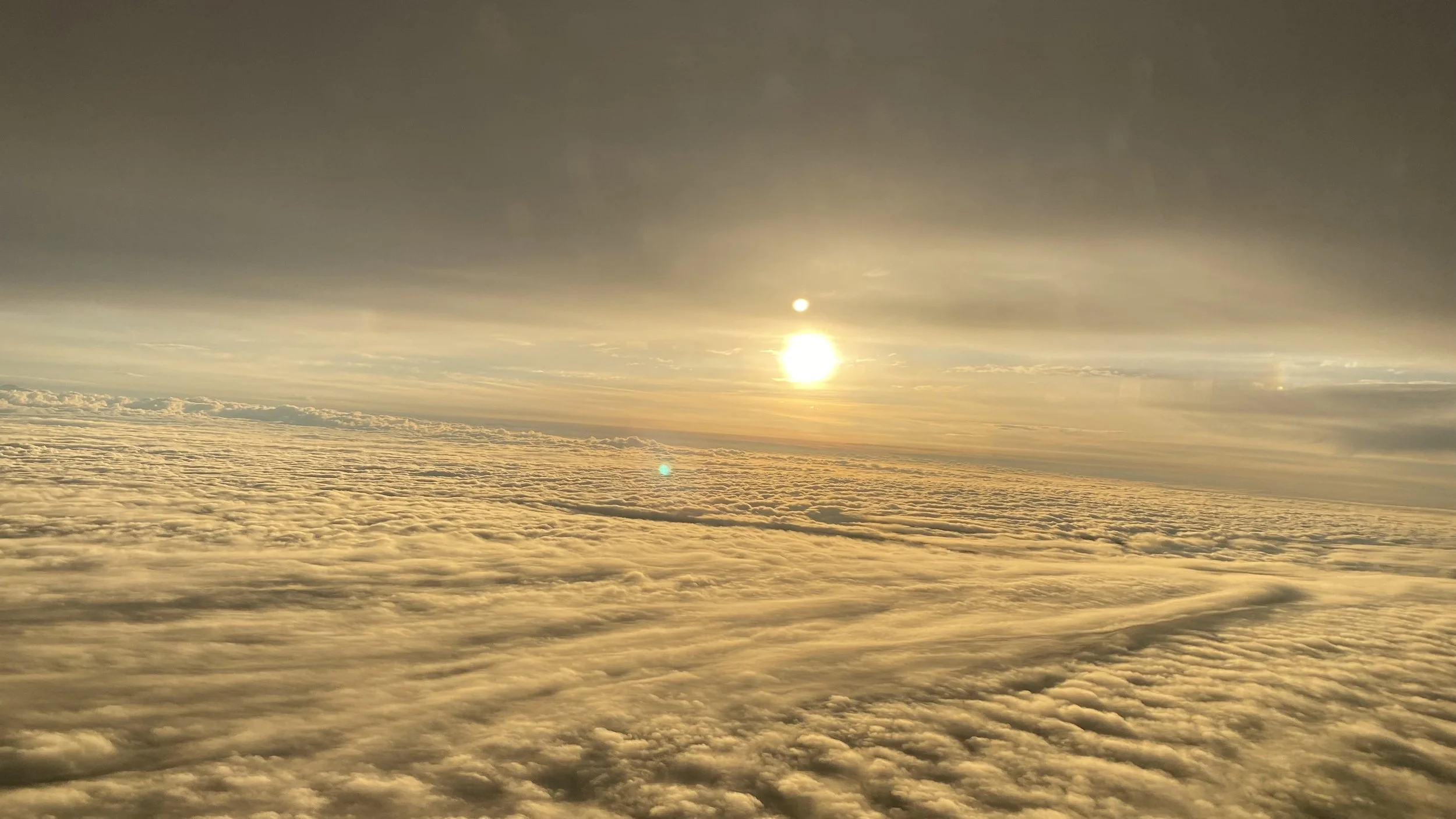 View above the clouds during sunset or sunrise with the sun glowing and a lens flare visible.