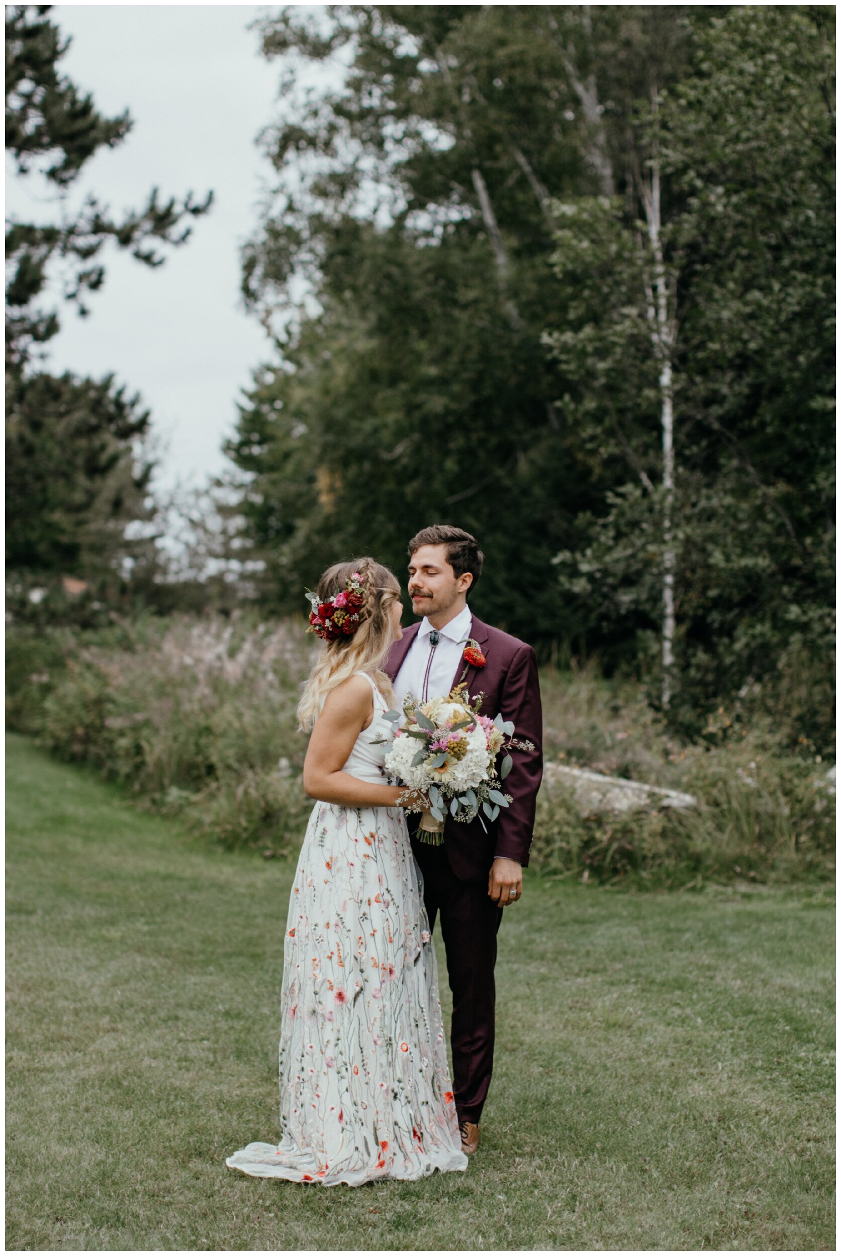 Bride wearing whimsical floral print wedding dress while looking at groom during Minnesota summer camp wedding in the woods