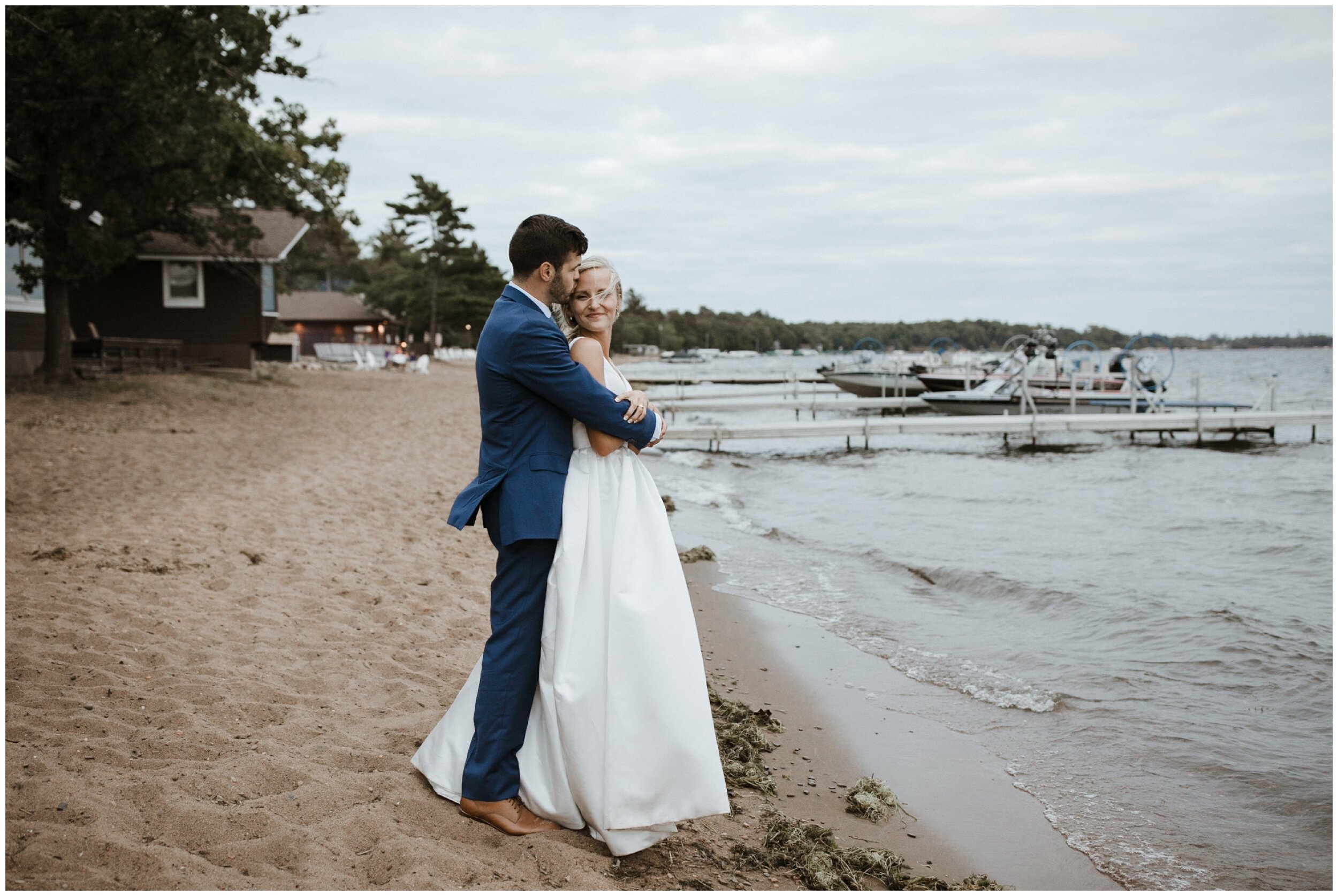 Groom kissing bride's forehead while standing on beach at Grand View Lodge wedding in Nisswa, Minnesota