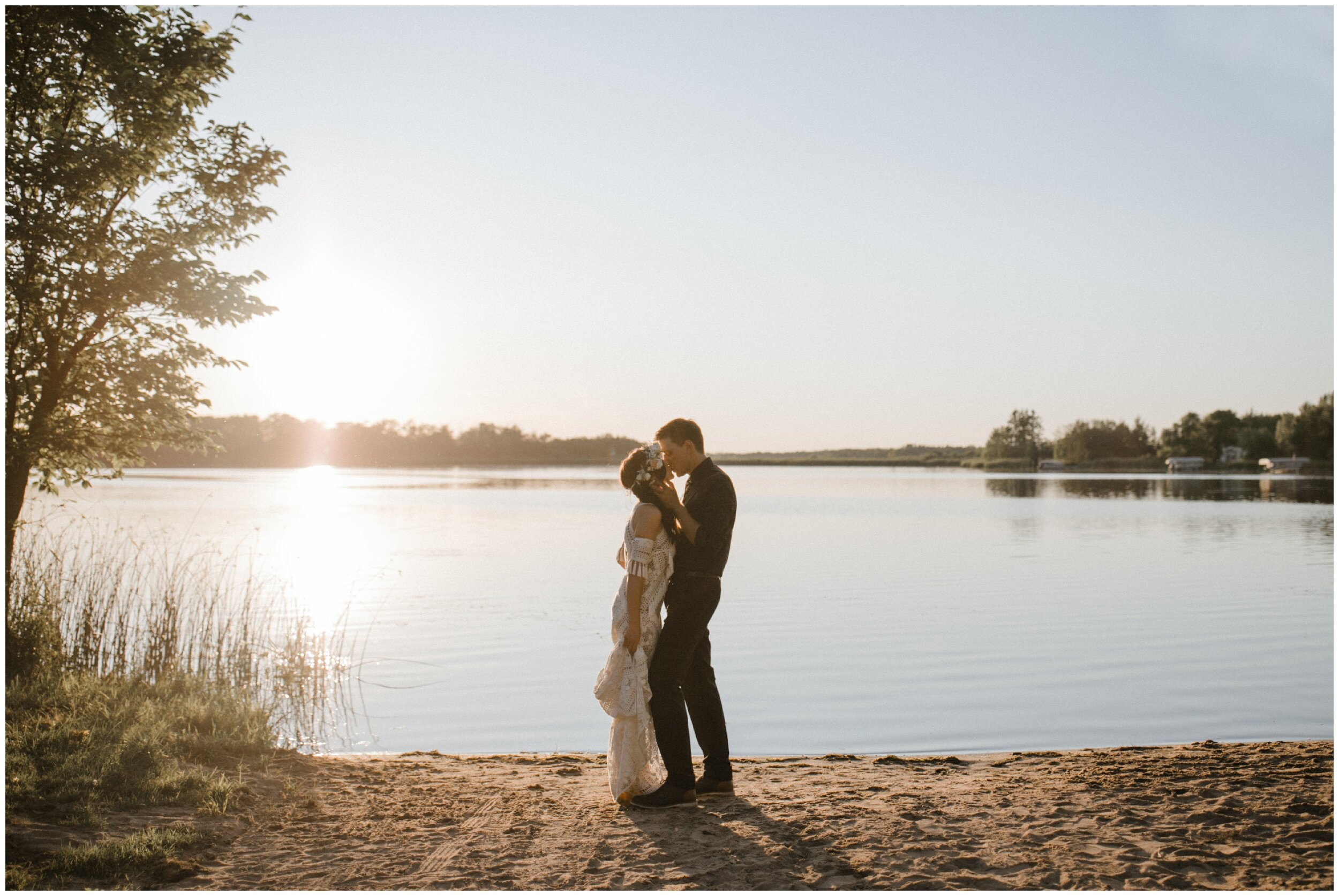 Bride and groom kissing while standing on lake beach during sunset at their Minnesota backyard summer wedding