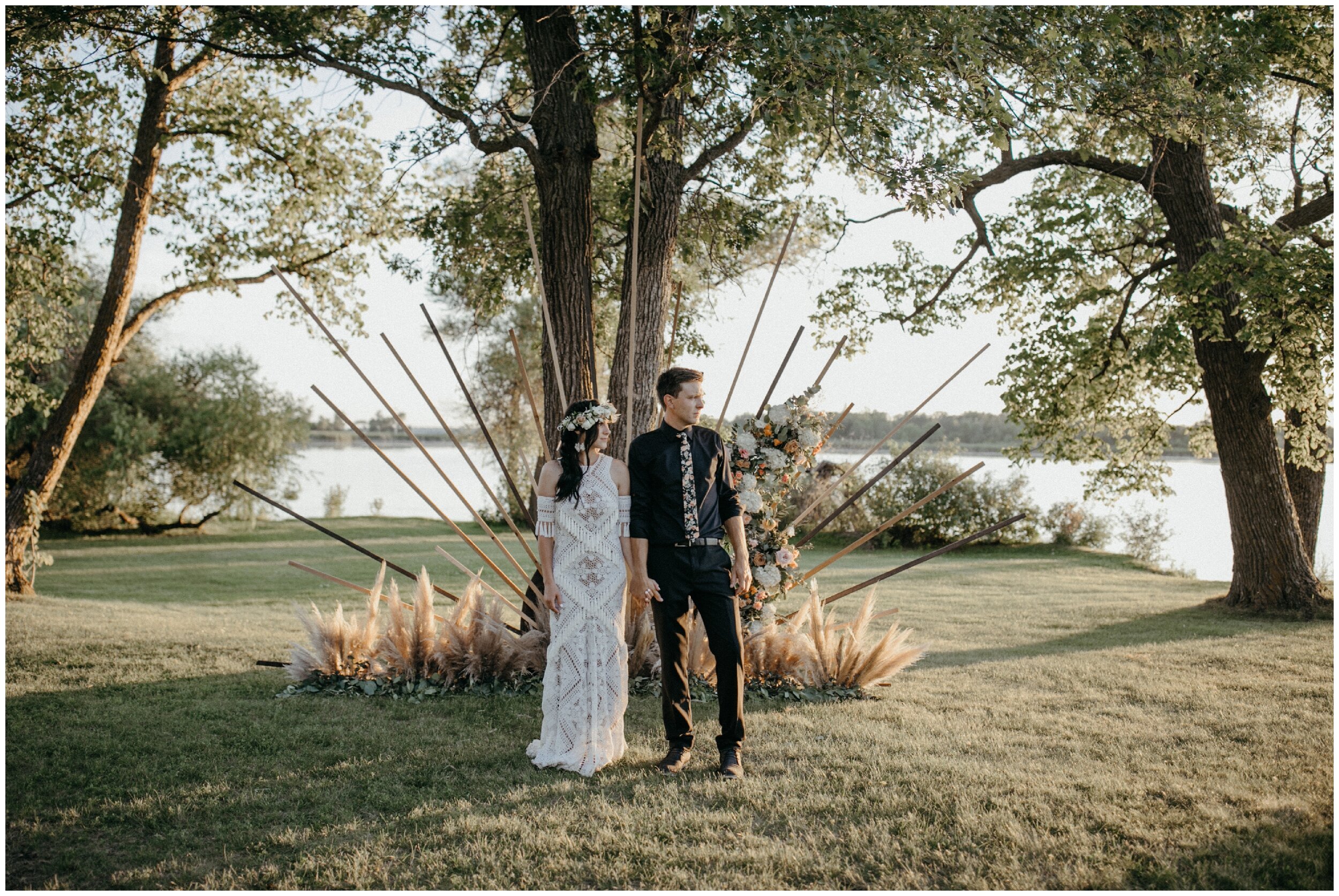 Bride and groom standing in front of boho themed wooden sunburst arch during sunset at their Minnesota summer backyard wedding