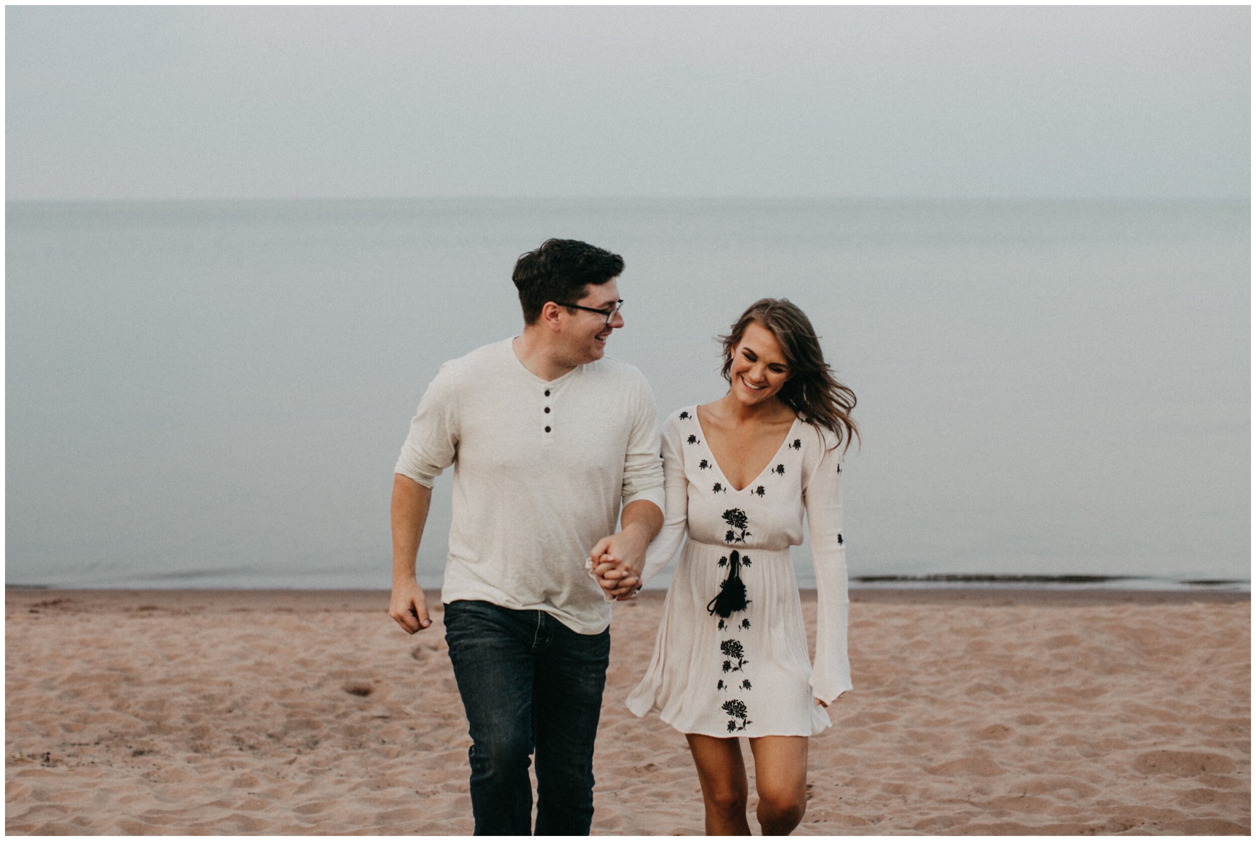 Couple laughing and walking on beach during engagement session at Park Point Beach in Duluth Minnesota