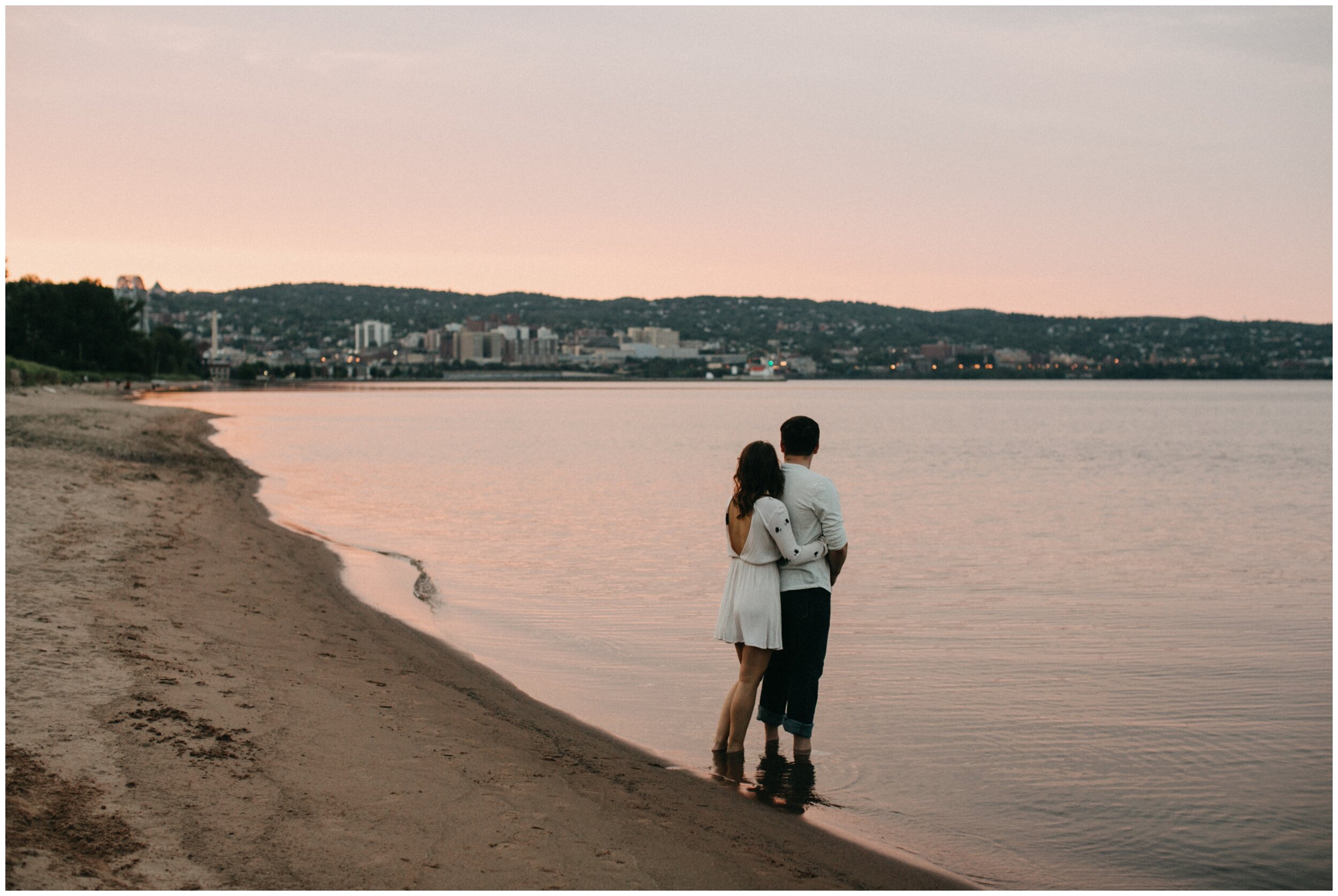 Couple standing along Lake Superior shore at Park Point Beach during engagement session in Duluth, Minnesota