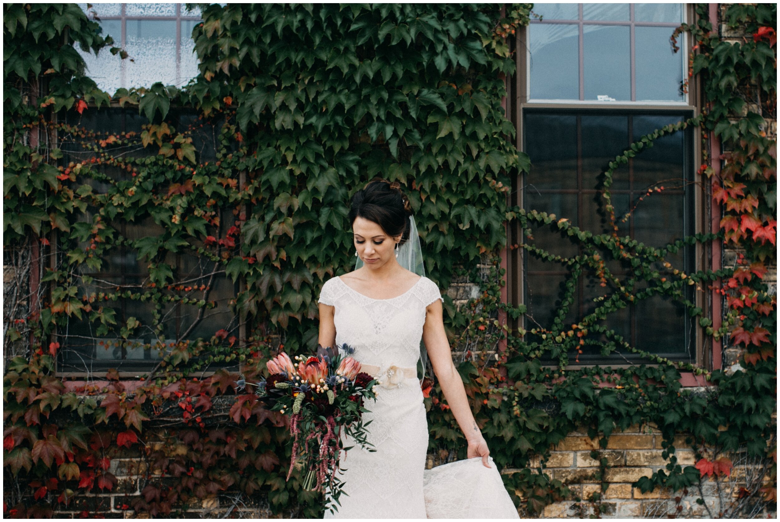 Bride looking at wedding bouquet while standing in front of ivy, covered brick wall at the Northern Pacific Center in Brainerd, MN