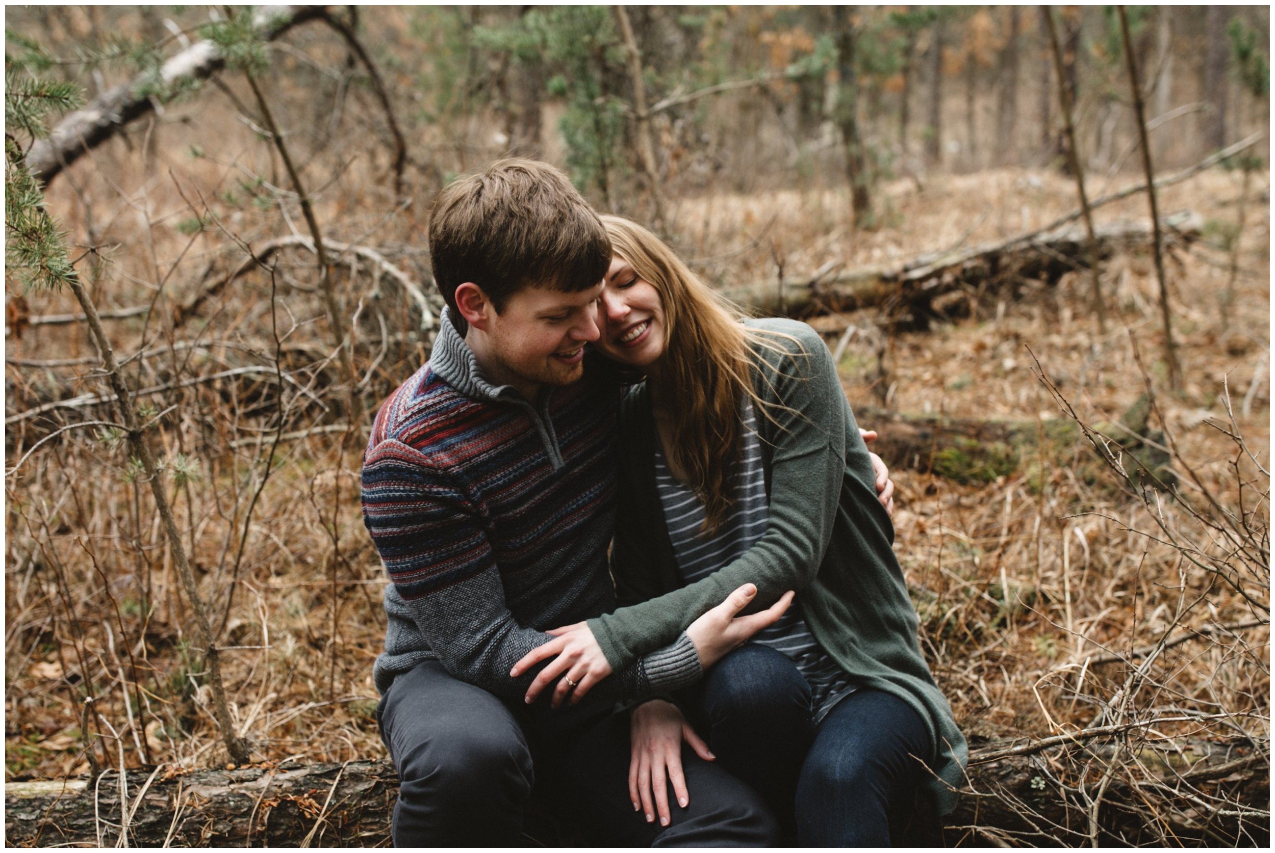 Couple sitting on log in the woods with arms wrapped around each other during Minnesota engagement session