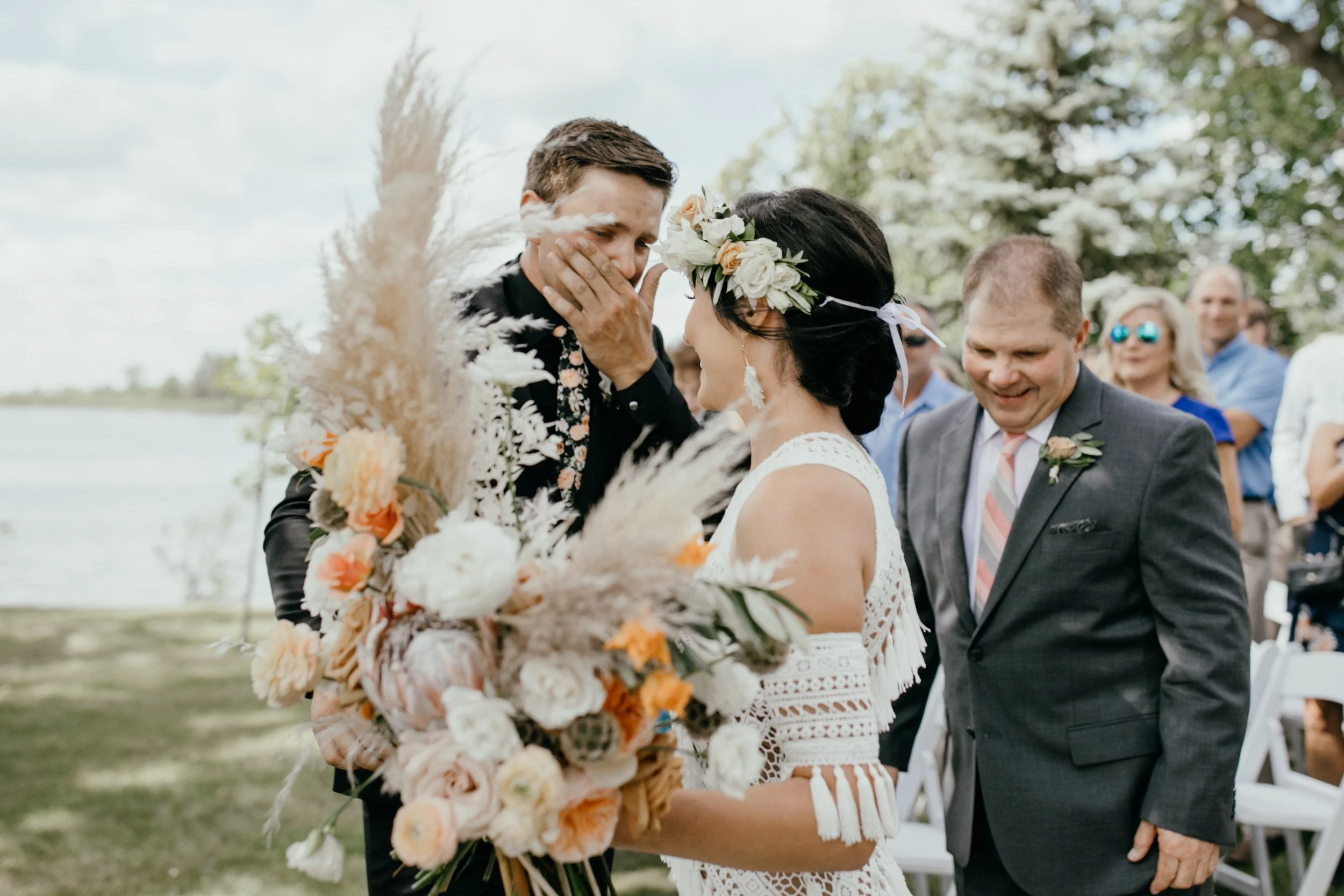 An emotional groom touching his face while see the bride walk dow the aisle, surrounded by friends and family at their backyard lakeside wedding ceremony in Minnesota.
