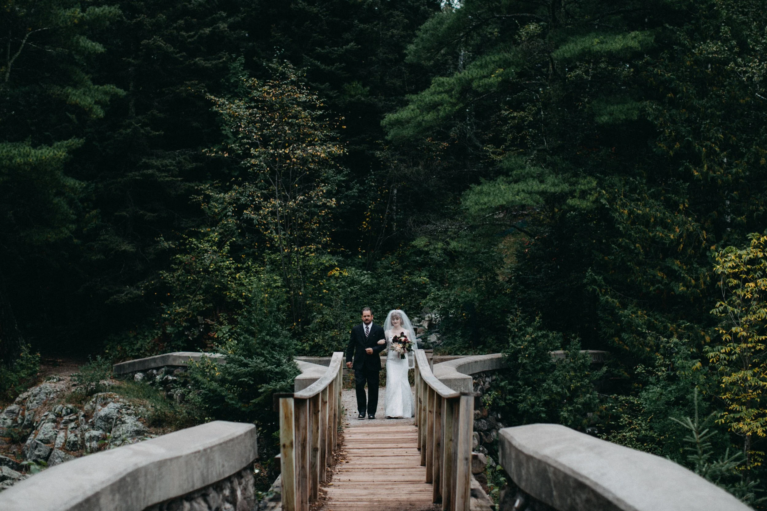 A bride and groom walking arm in arm on a small wooden bridge in a lush green forest during a wedding in Minnesota.
