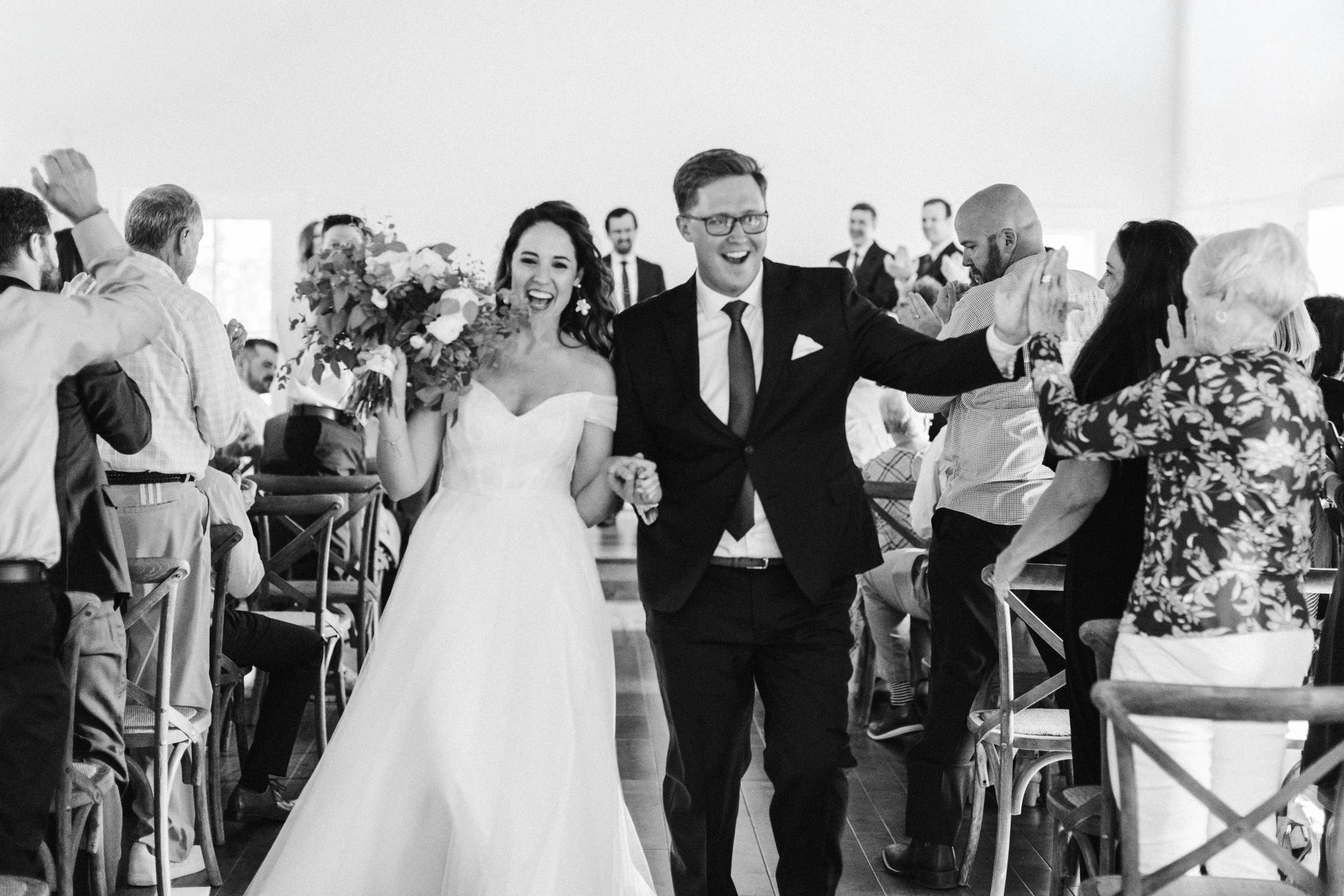 A bride and groom walking down the aisle, smiling and holding hands after their wedding ceremony in the white chapel in the woods at Grand View Lodge in Nisswa Minnesota, surrounded by seated guests applauding and celebrating.
