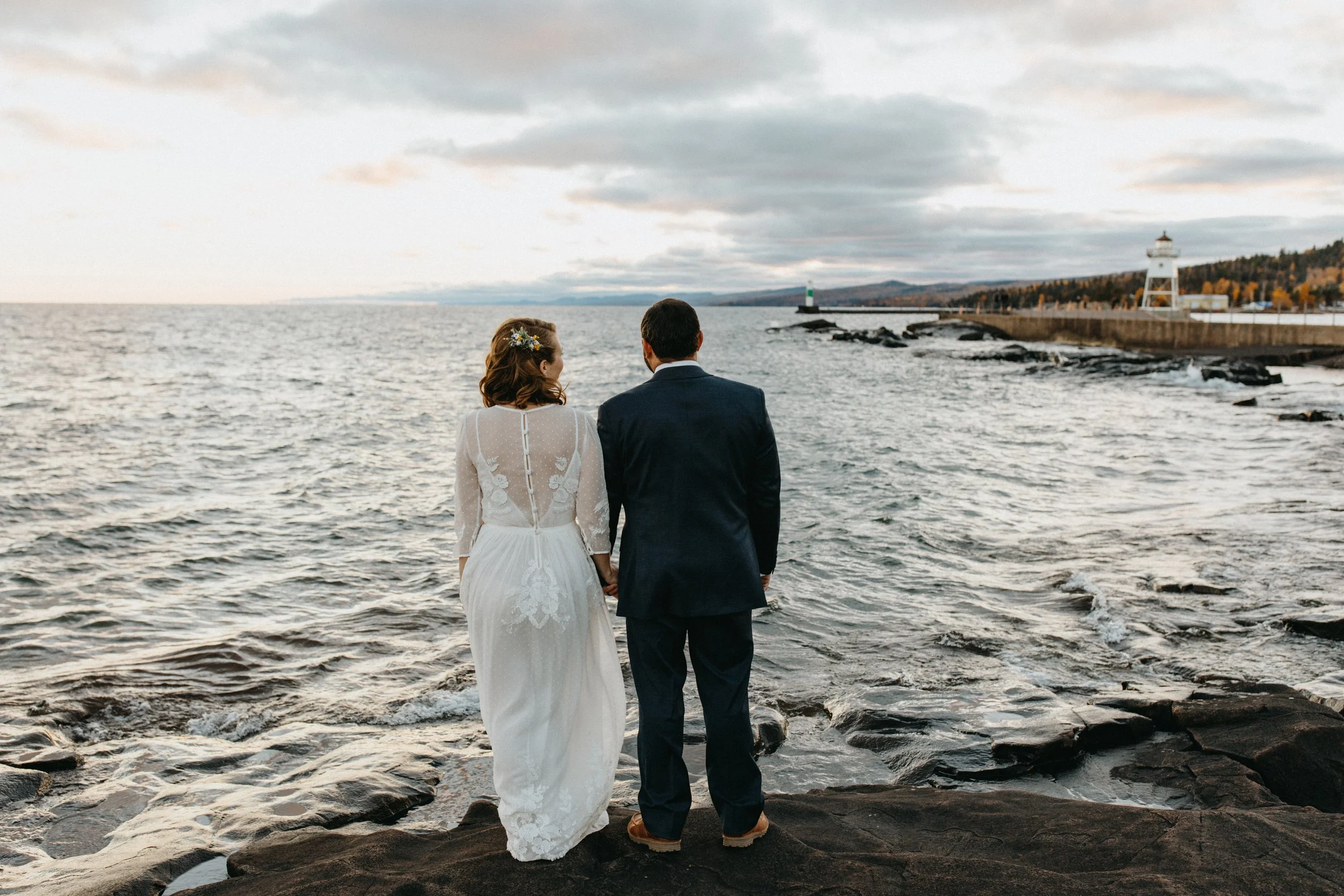 Bride and Groom standing on rocky shore looking out at lighthouse of Artist's Point in Grand Marais Minnesota