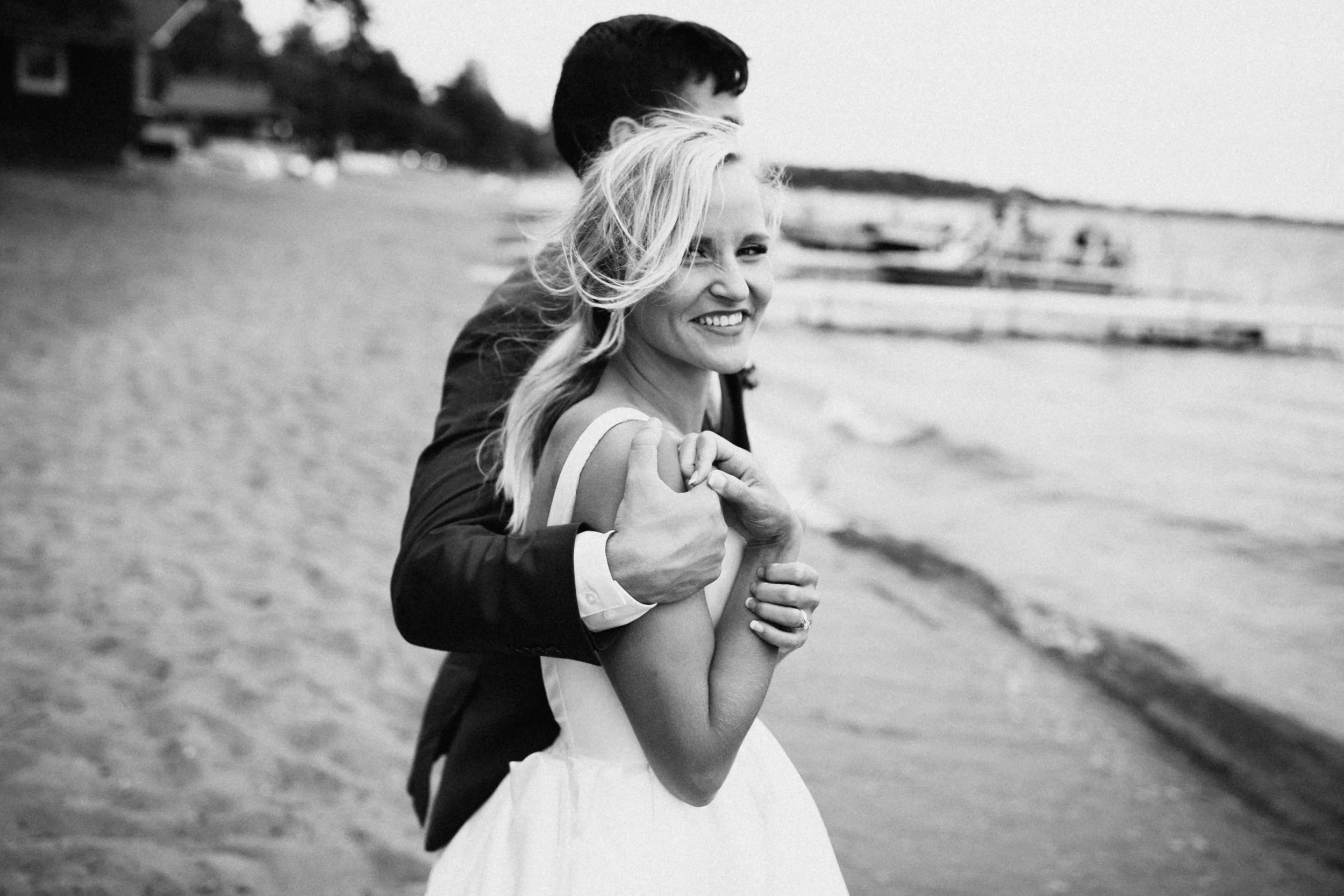Bride and groom standing on beach with arms wrapped around each other. The groom is look out at Gull Lake and the bride is smiling at the camera.