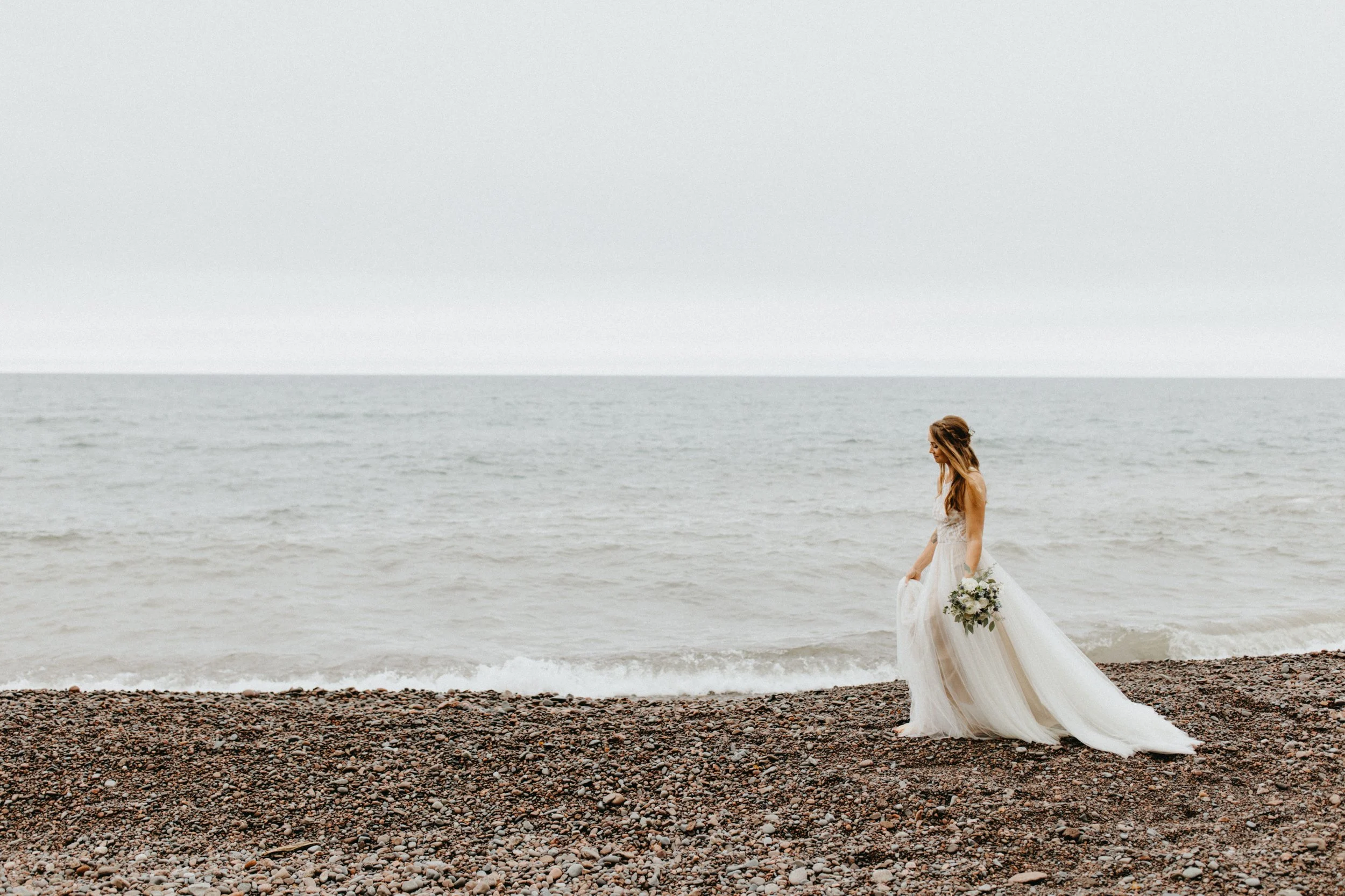 Bride wearing a flowy tulle wedding dress walking along rocky Lake Superior beach in Lutsen, Minnesota
