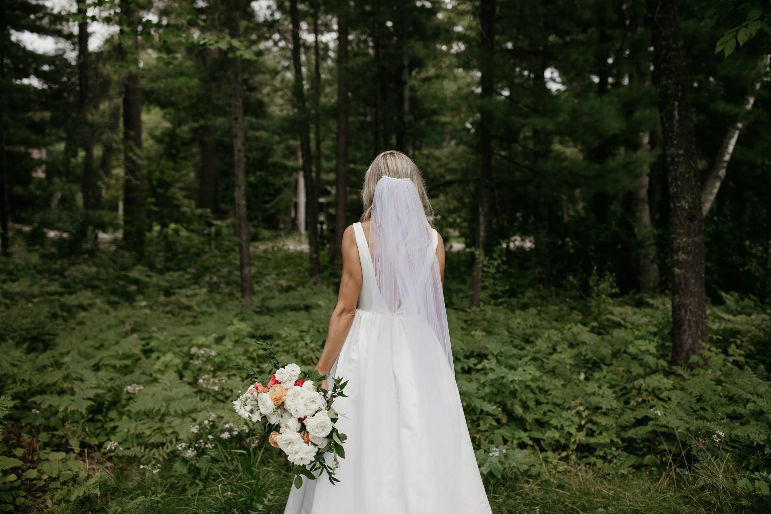 Bride wearing cathedral veil standing in front of lush green forest on her wedding day in Minnesota