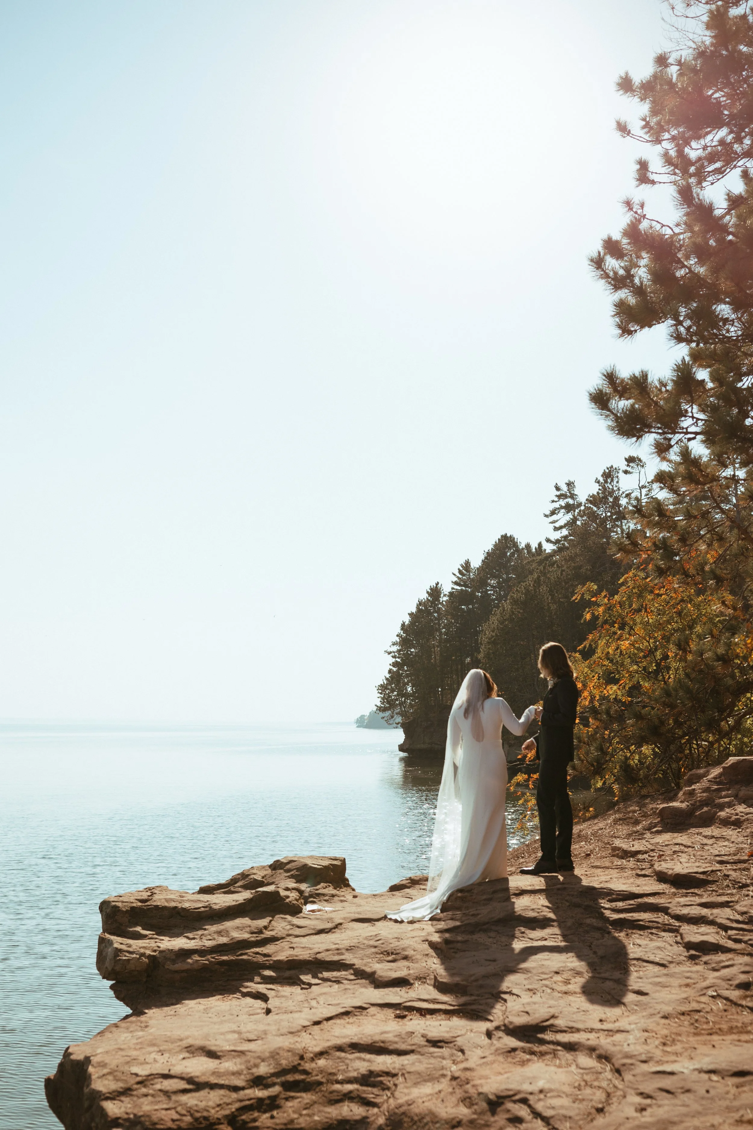 Bride and groom holding hands on a rocky lakeshore with fall trees, photographed by a Stillwater, Minnesota wedding photographer.