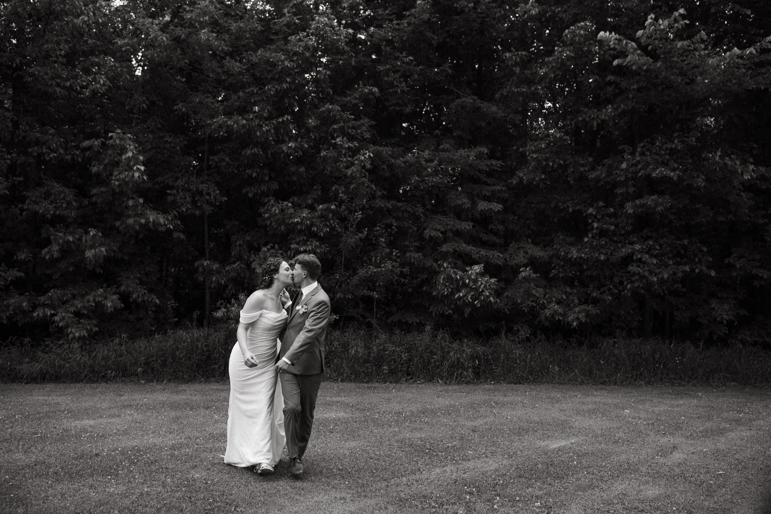 Bride and groom sharing a kiss as they walk across a grassy field in front of a line of trees photographed by St. Paul, Minnesota wedding photographer Britt DeZeeuw.