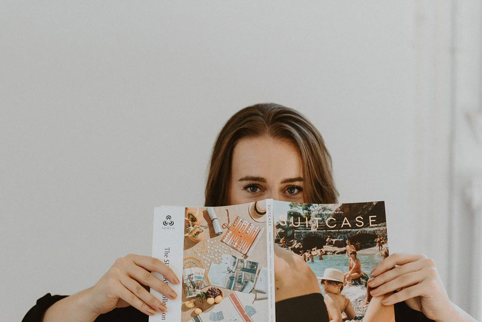A woman with brown hair holding a colorful travel magazine, partially hiding her face, looking over the magazine with blue eyes, and a plain white wall background.