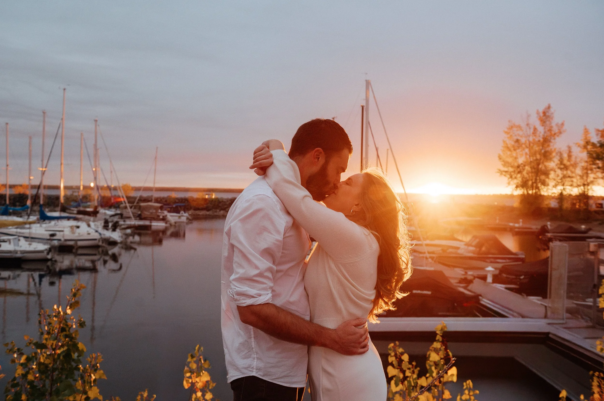 A couple kissing at sunset near a marina with boats and sailboats docked in the background.