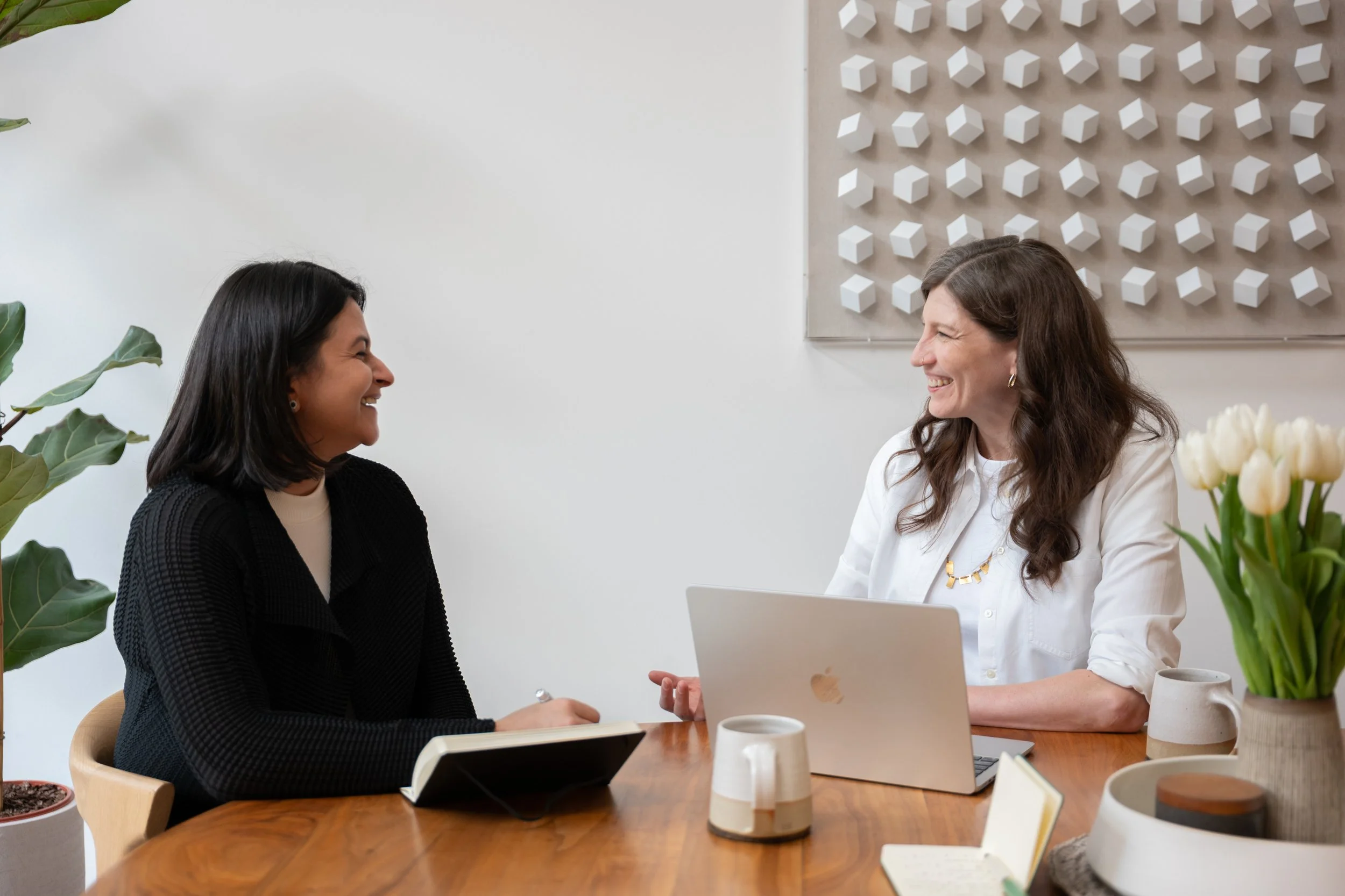 Two women are sitting at a table and laughing; there is a laptop and a notebook on the table between them