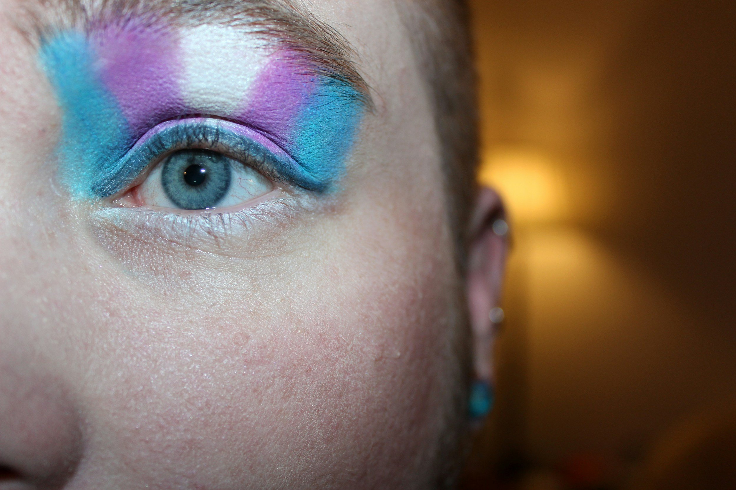 A person with pale skin and a shaved head wears eyeshadow in the colours of the trans pride flag