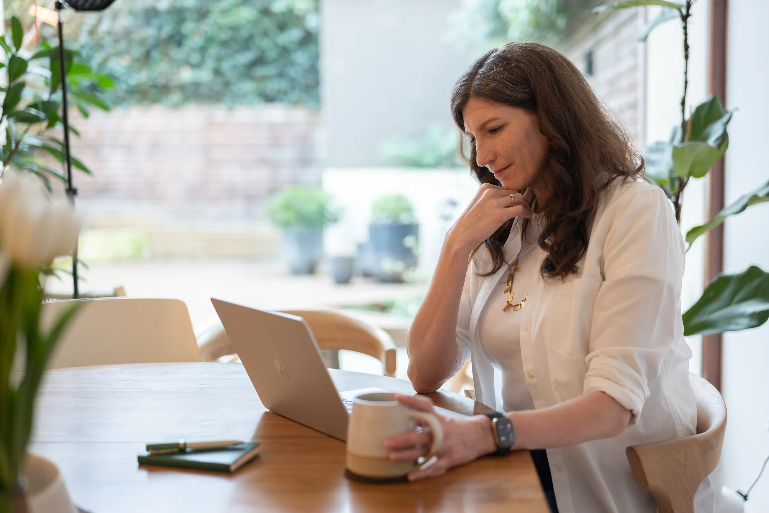 A woman looks at a laptop screen, she holds a mug.