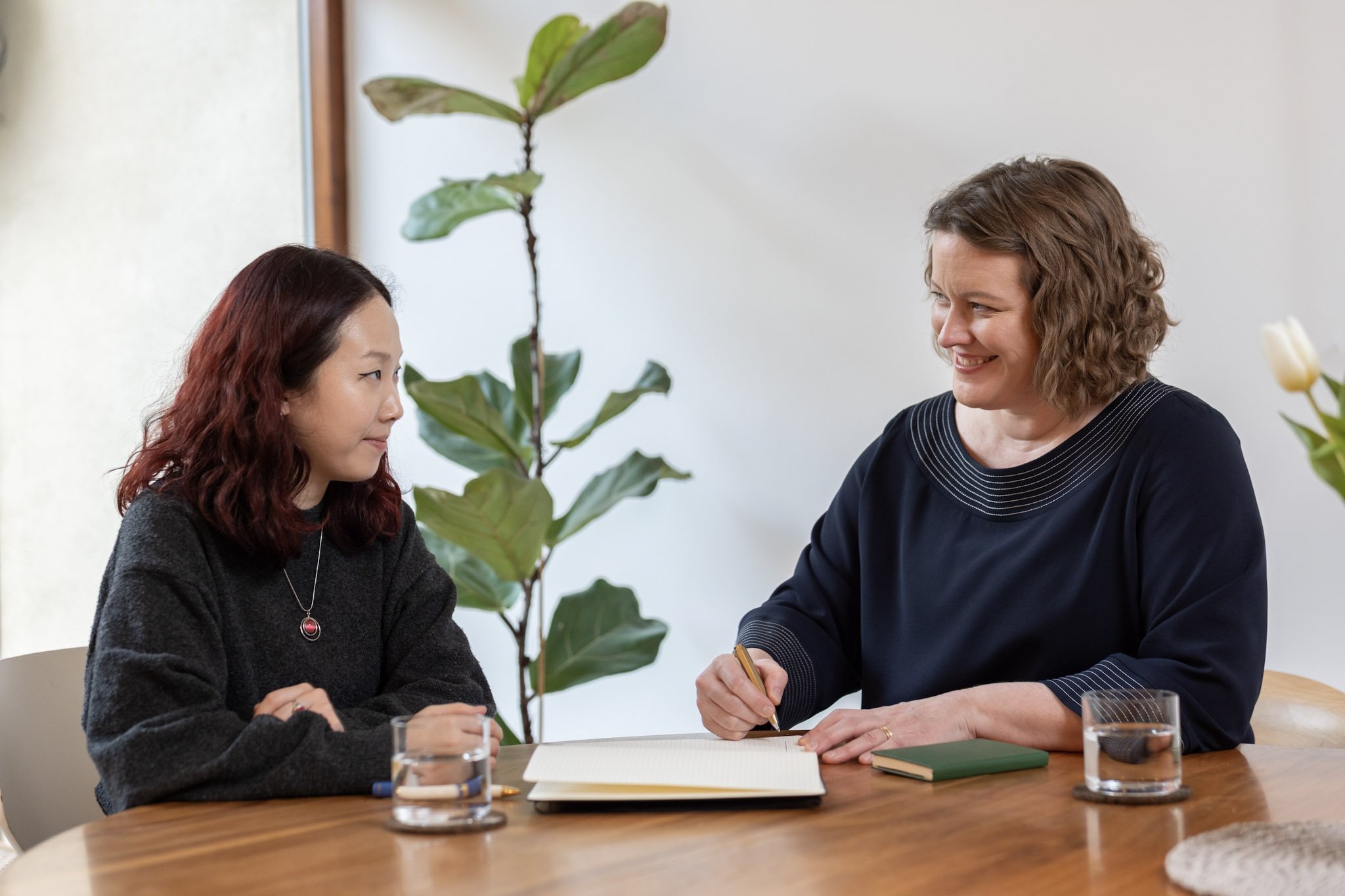 Two women are sitting at a table with a notebook open between them.