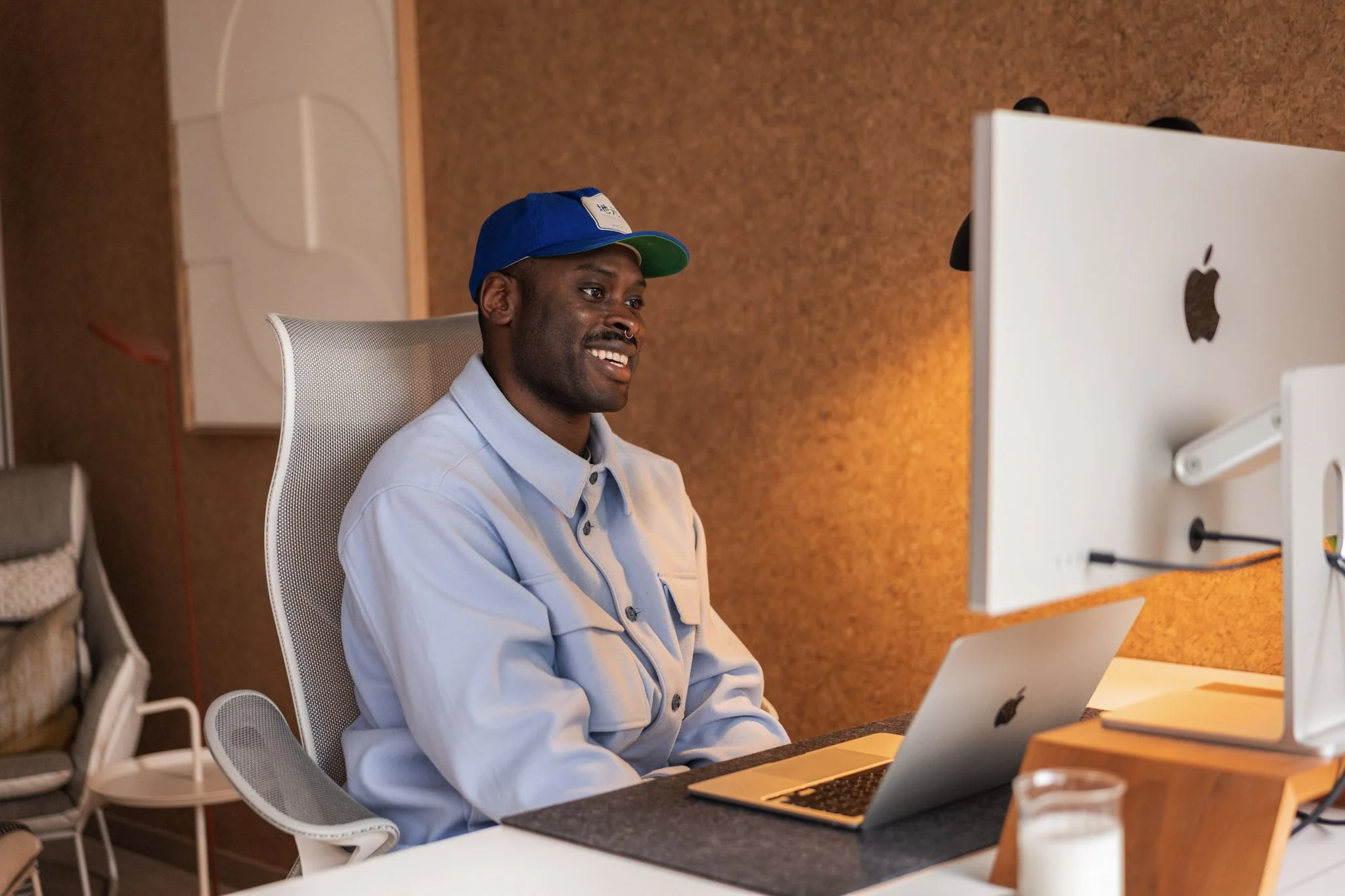 A man with dark skin wearing a pale blue shirt and blue baseball cap smiles at a computer screen, on a video call