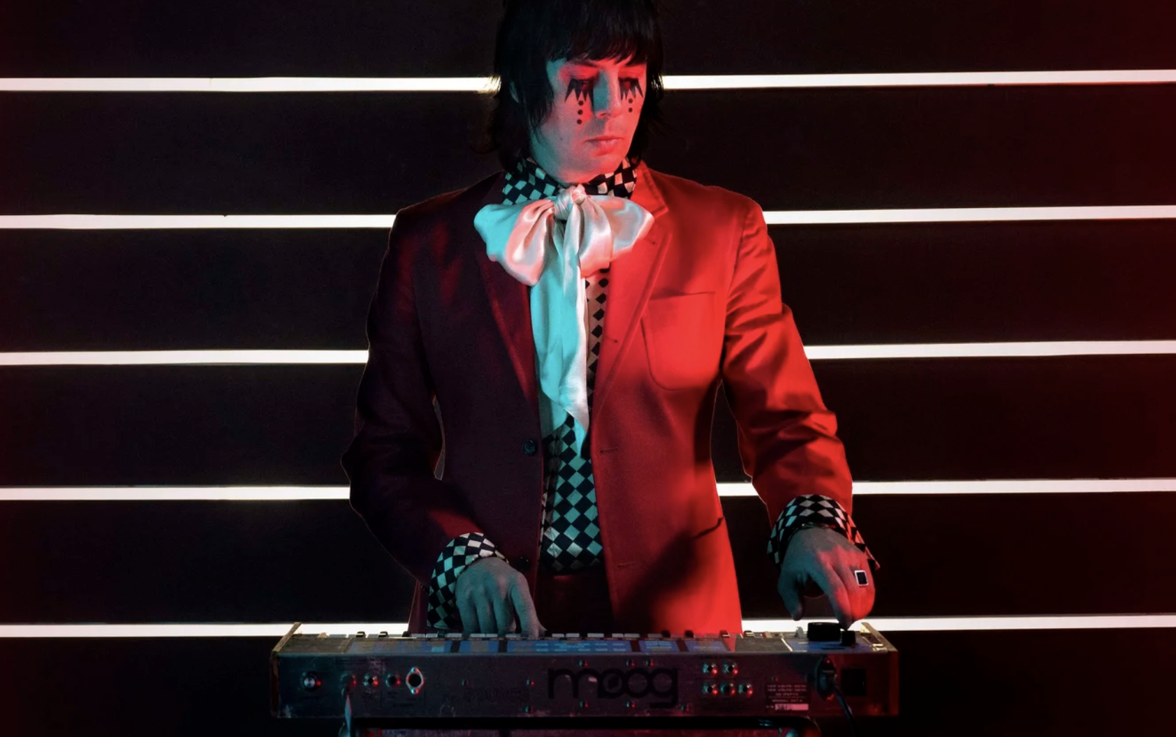 Johnny Jewel stands behind a keyboard, wearing a black and white harlequin patterned shirt with a red jacket and a large neck bow.