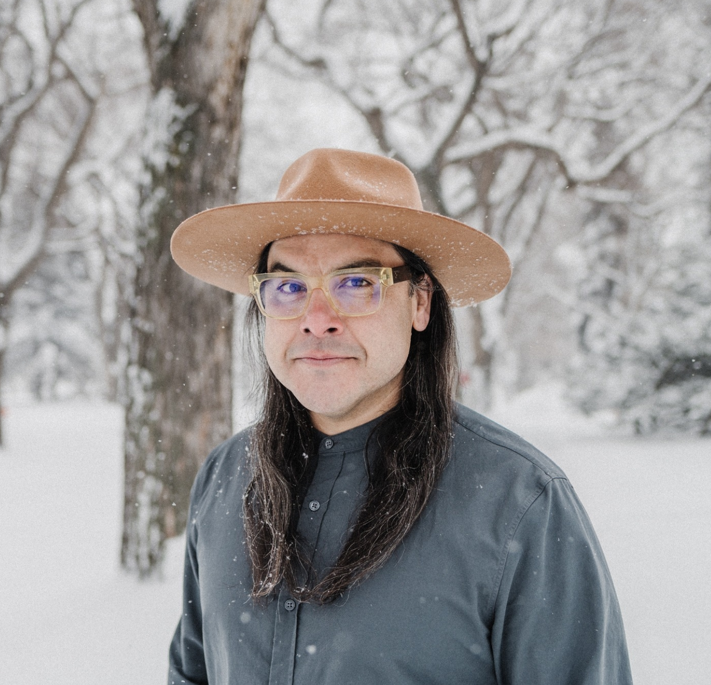 Marek Tyler stands in a snowy wooded area wearing a button up shirt and a wide brimmed hat and glasses.
