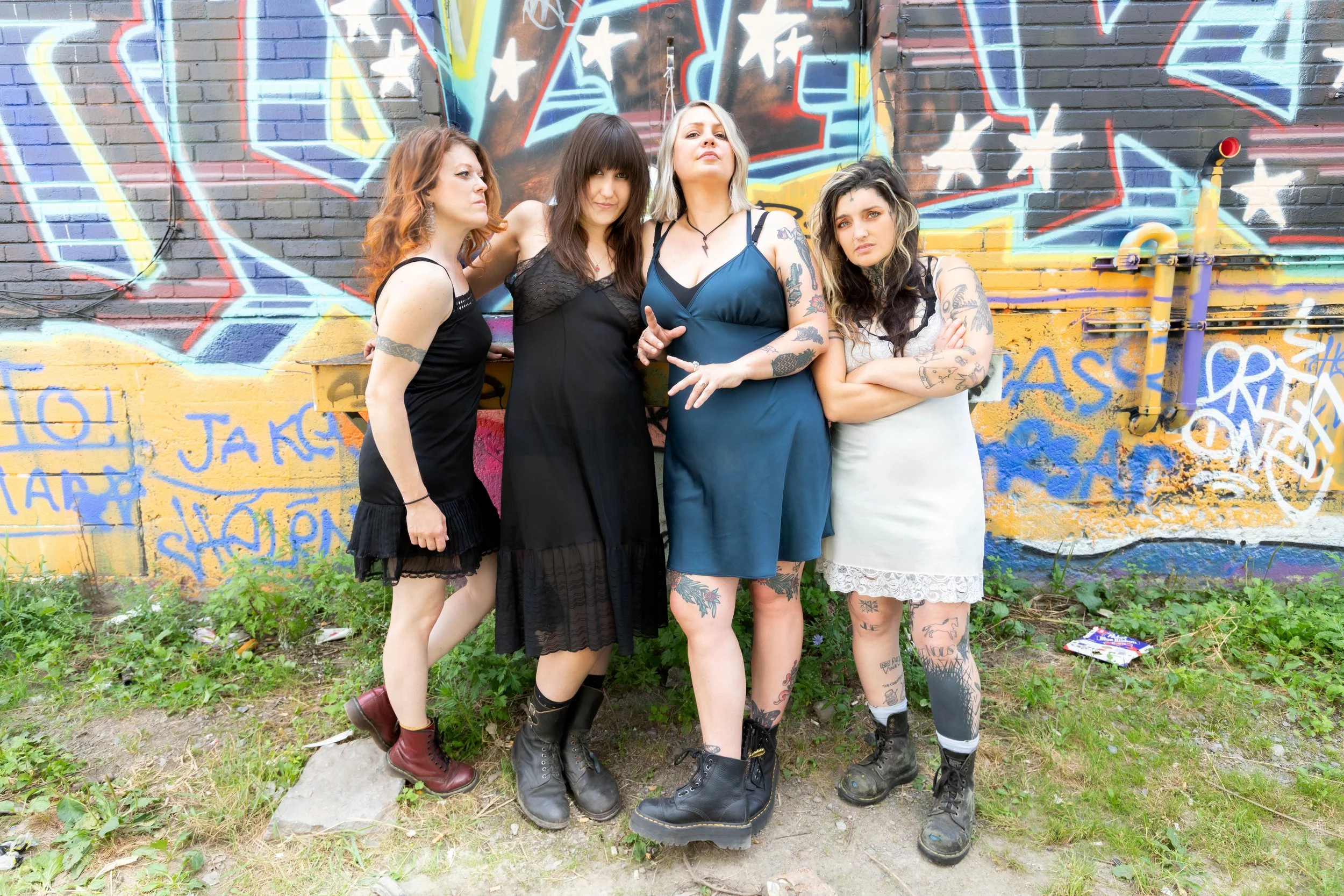 Taxi Girls wear slip dresses and combat boots, standing in front of a graffitied wall.