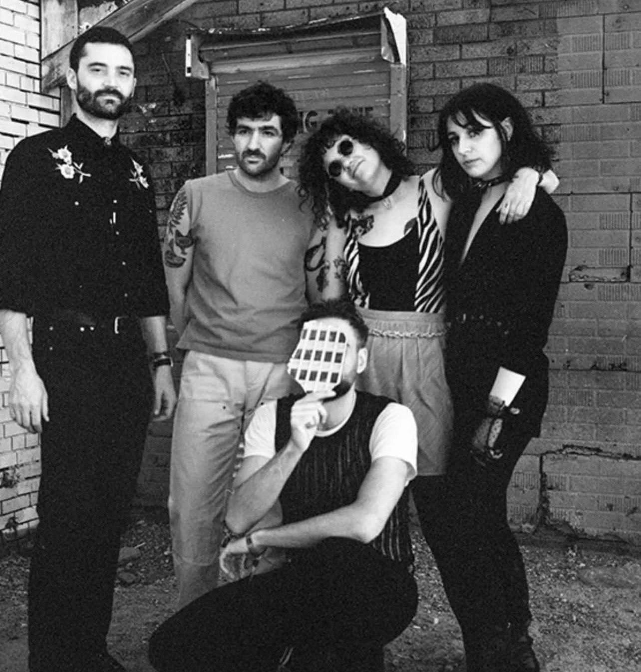 The five members of Uncanny Valley stand against a brick wall and a roll-down door.