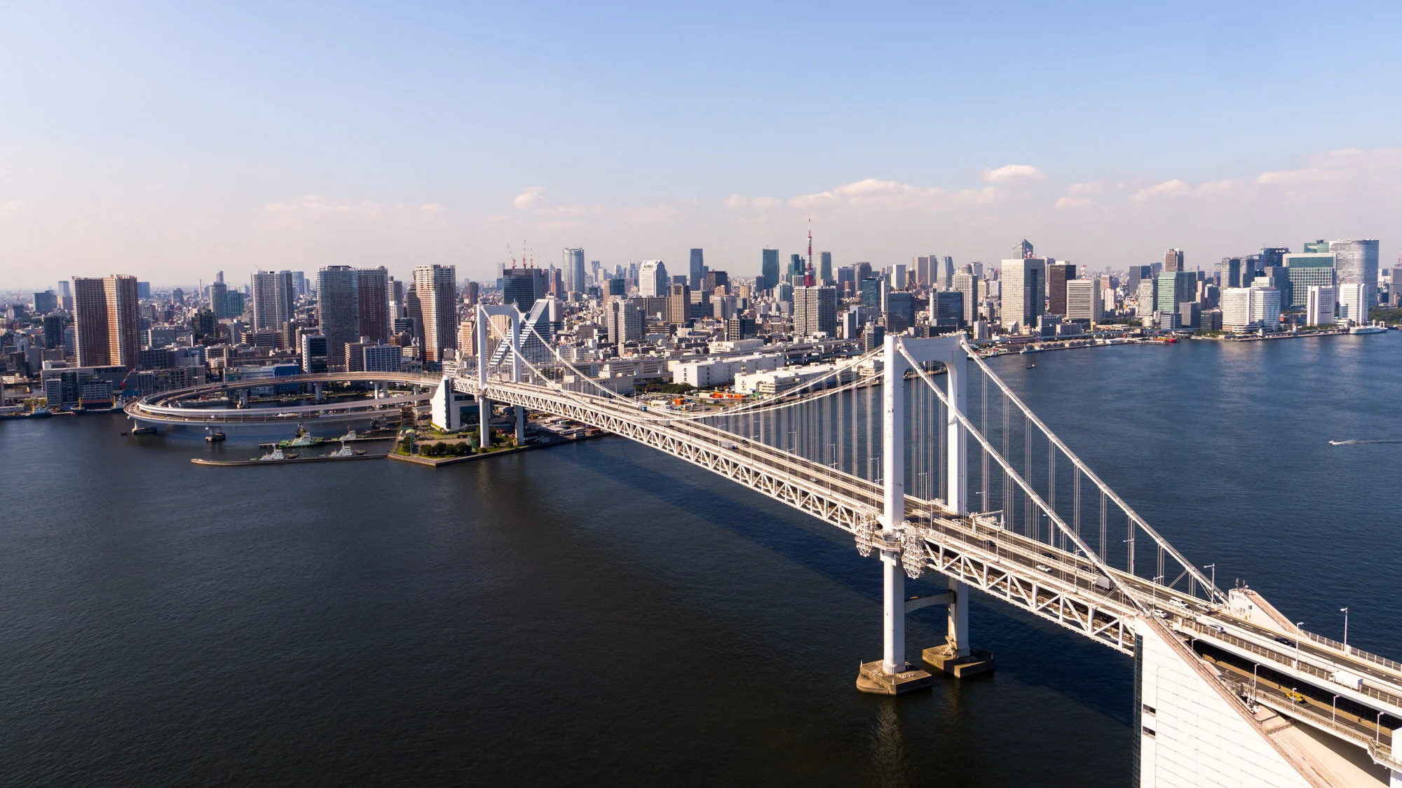 Aerial view of a city skyline with a large suspension bridge over a body of water, featuring numerous skyscrapers and buildings under a partly cloudy sky.
