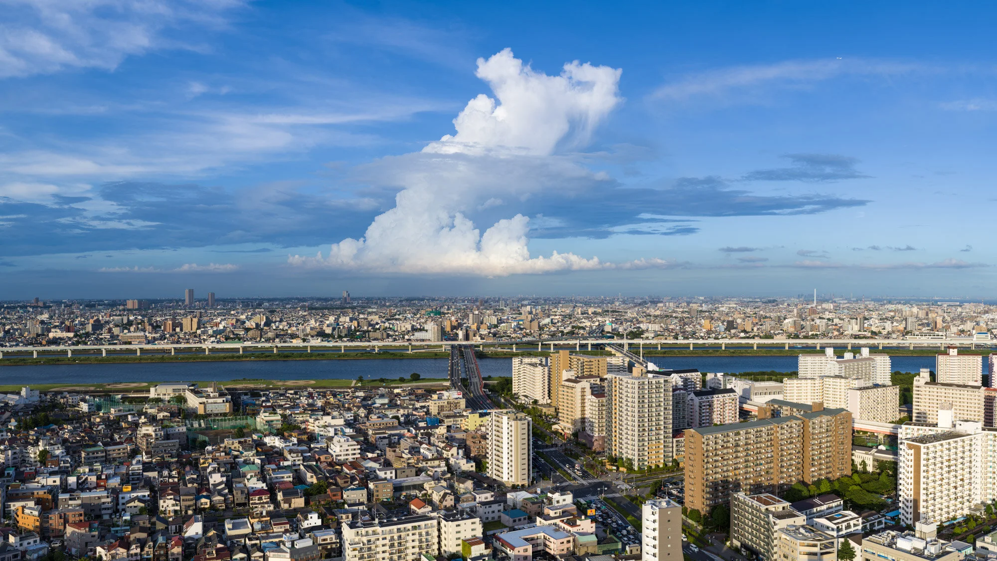 Aerial view of a cityscape with high-rise buildings, bridges, water bodies, and a large cloud formation in the sky.