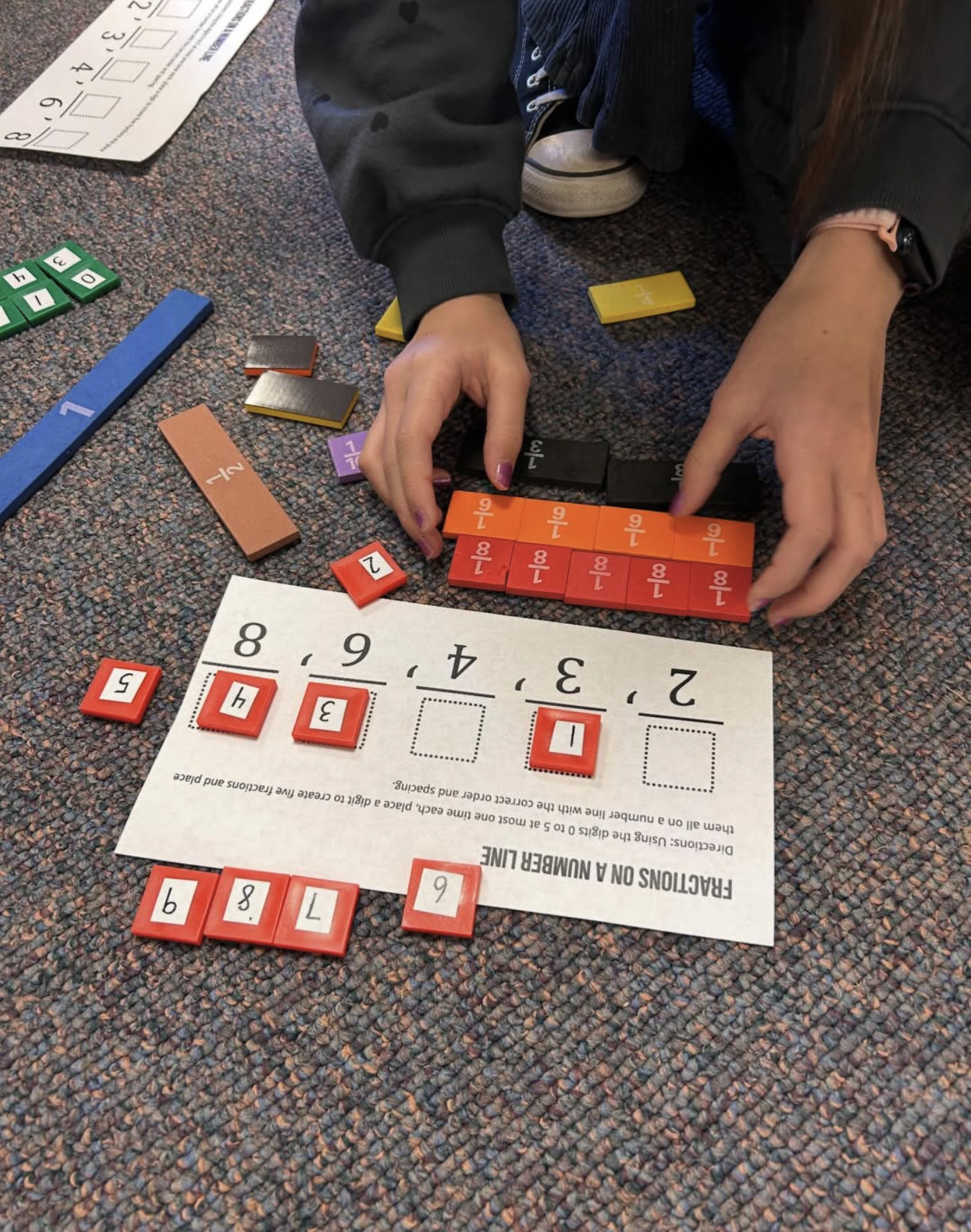 A person assembling a math puzzle on a carpeted floor, with colorful tiles and a worksheet with fractions and numbers.