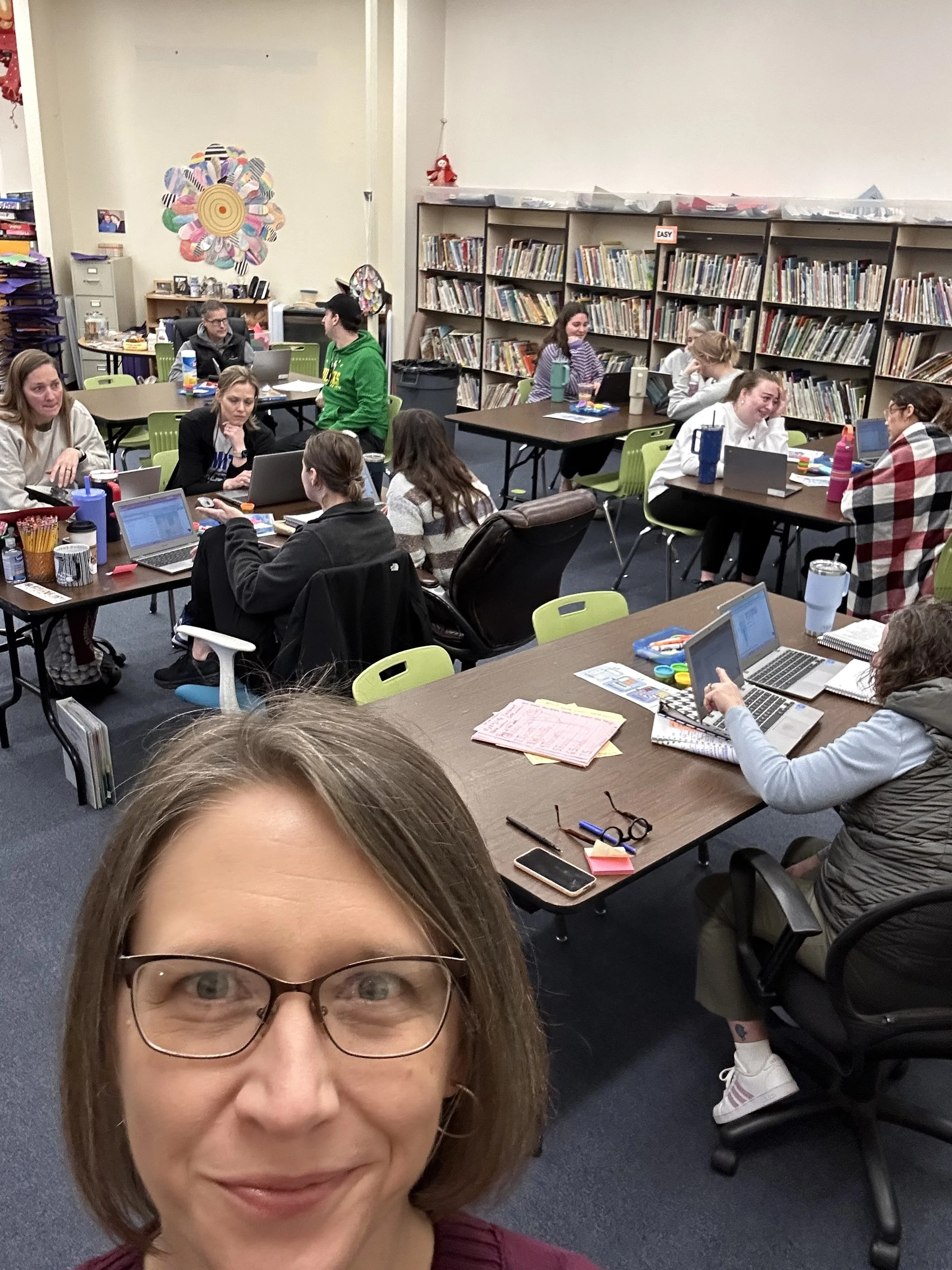 A woman with glasses taking a selfie in a classroom with students working at tables in the background.