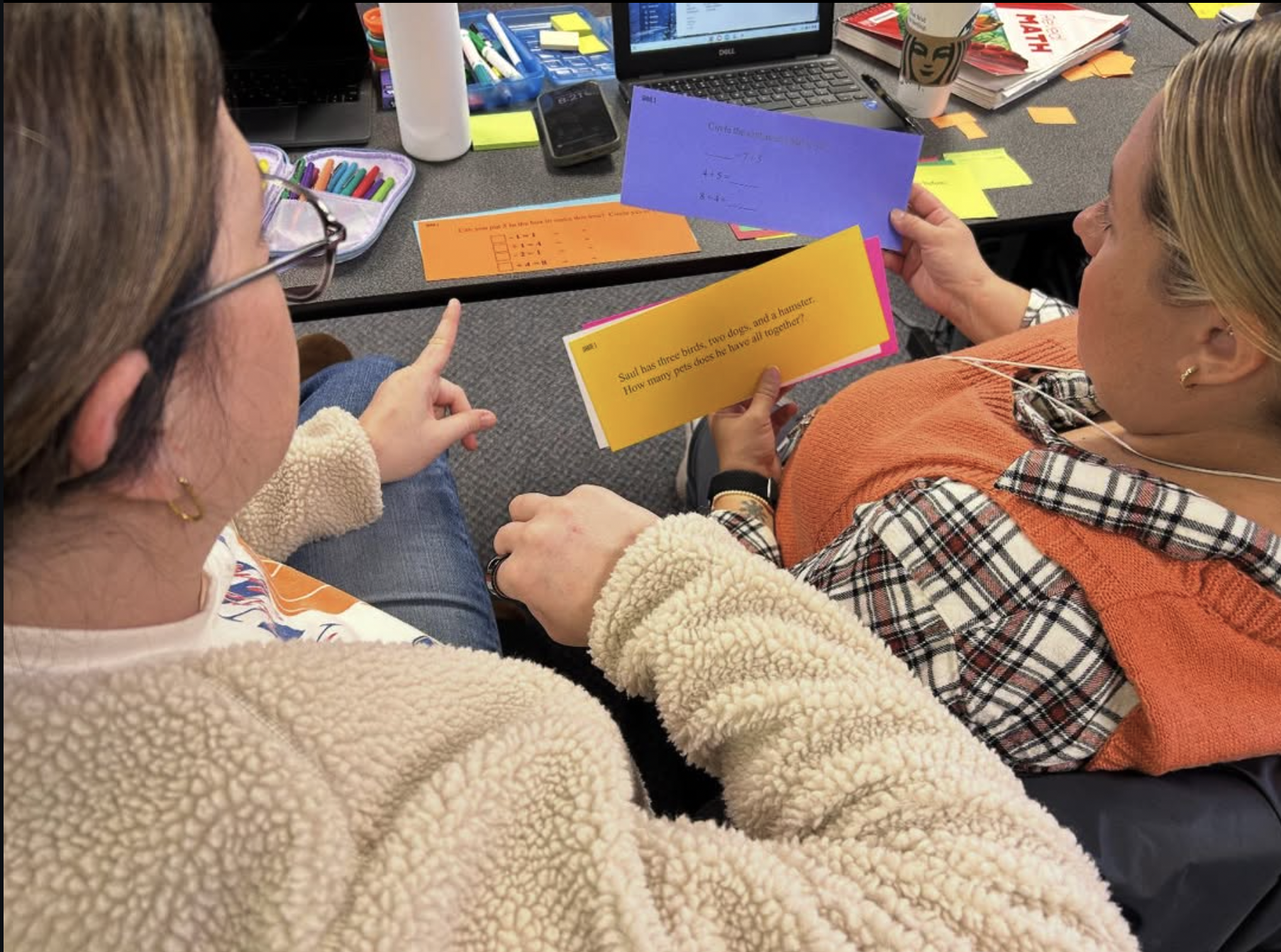 Two women sitting at a table engaged in a learning activity, with one woman holding an educational card and the other pointing at it. The table is cluttered with school supplies, a laptop, and books.