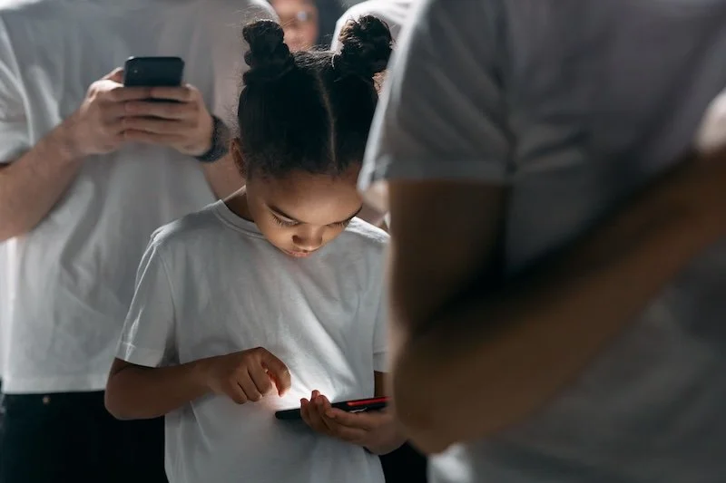 Enfant sur un portable au milieu d'adultes également sur leurs portables. IA et enfants, quels risques pour les apprentissages...