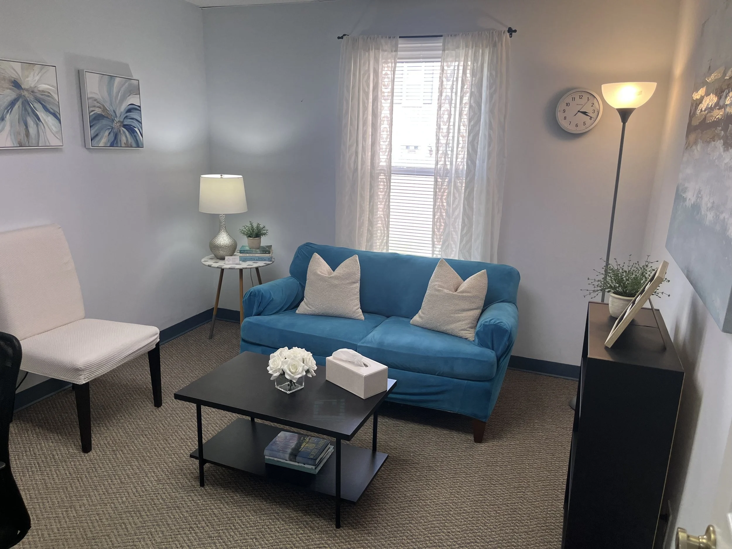 A cozy living room with a blue velvet sofa, white pillows, a black coffee table with a flower arrangement and books, a white armchair, a side table with a lamp and a plant, a window with sheer curtains, wall art, a clock, and a floor lamp.